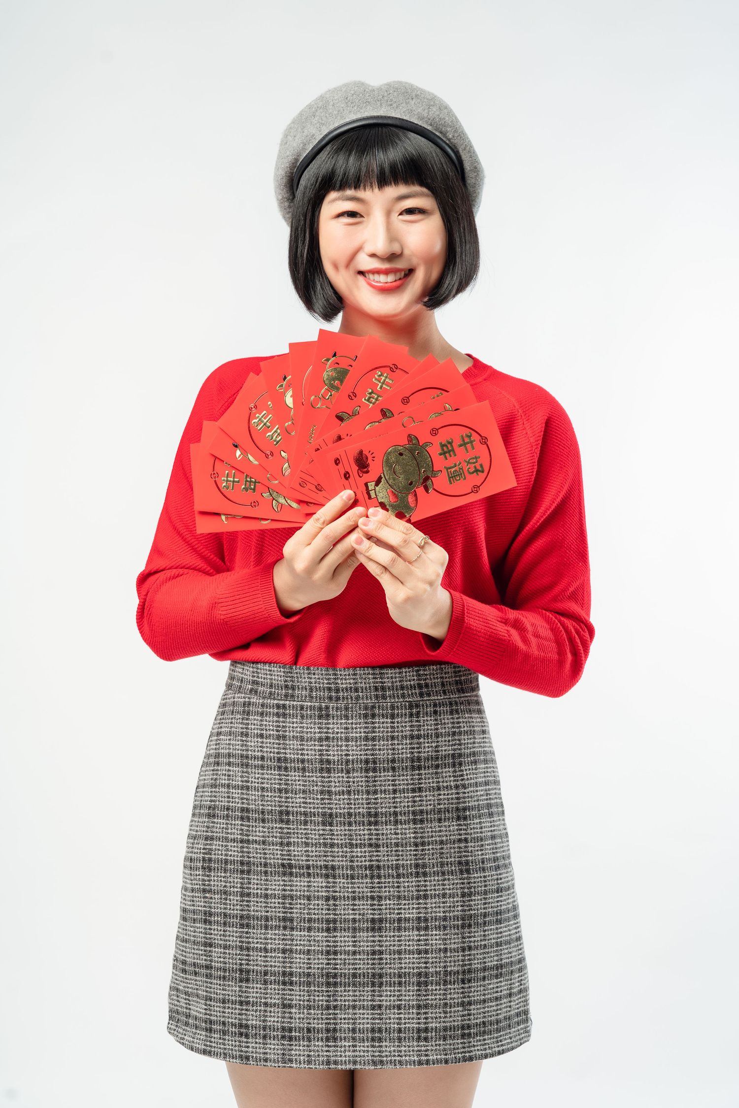 Young woman in red sweater, plaid skirt and beret holding traditional Chinese red envelopes against white background.
