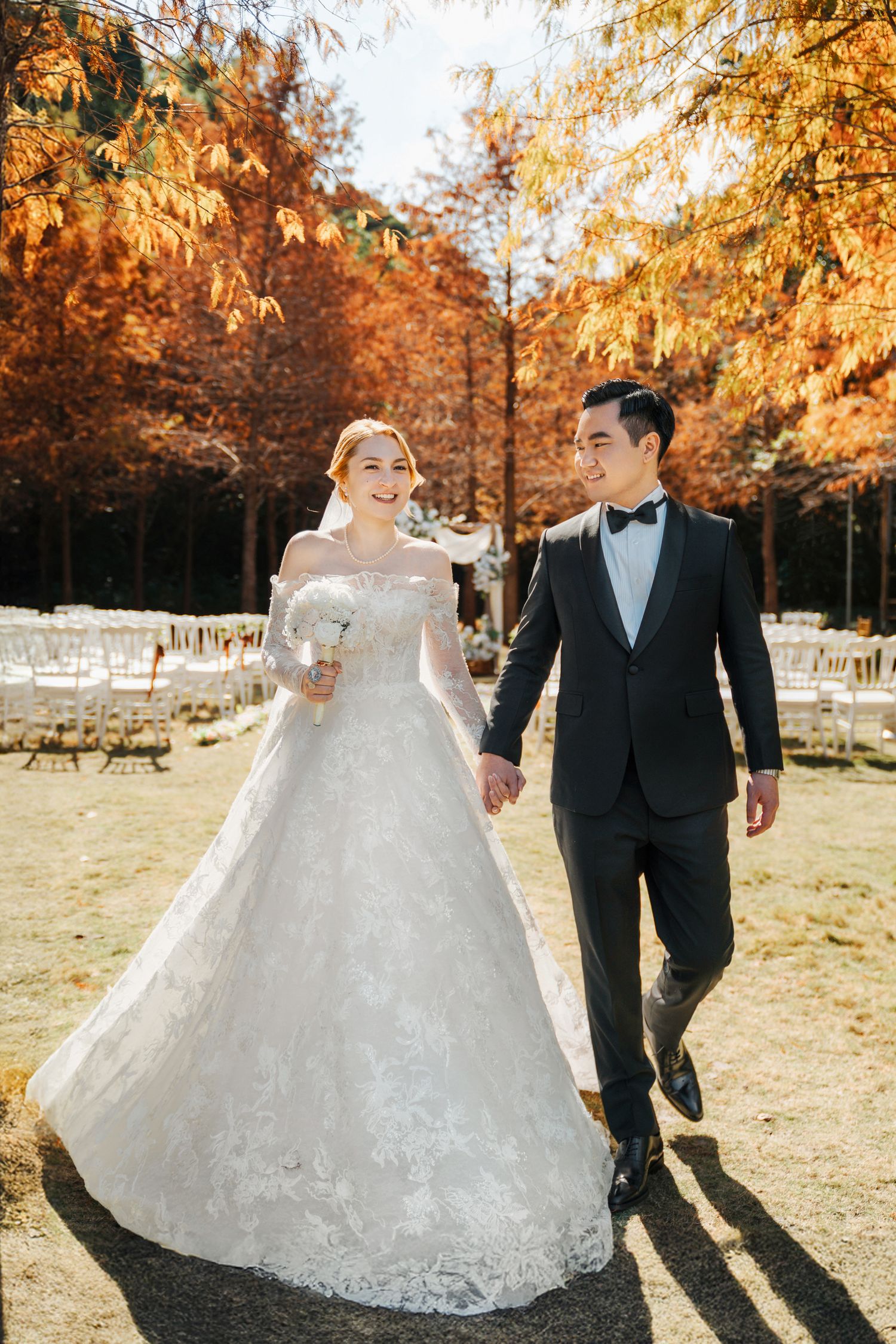 A couple in formal wedding attire walks hand in hand among autumn trees with golden foliage in an outdoor ceremony setting.