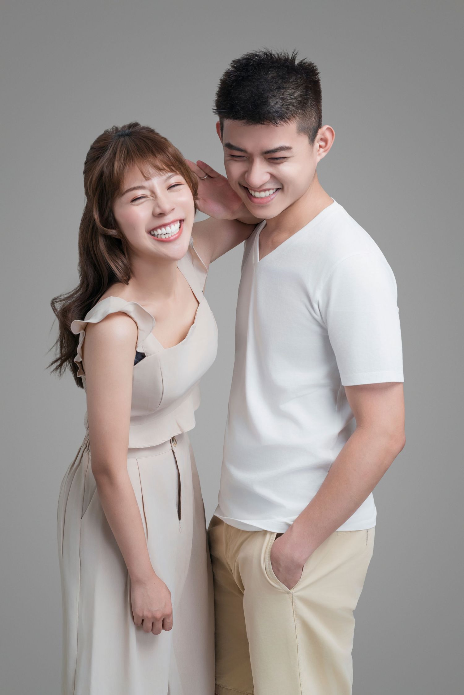 A couple in white and beige clothing share a joyful moment together against a neutral background.