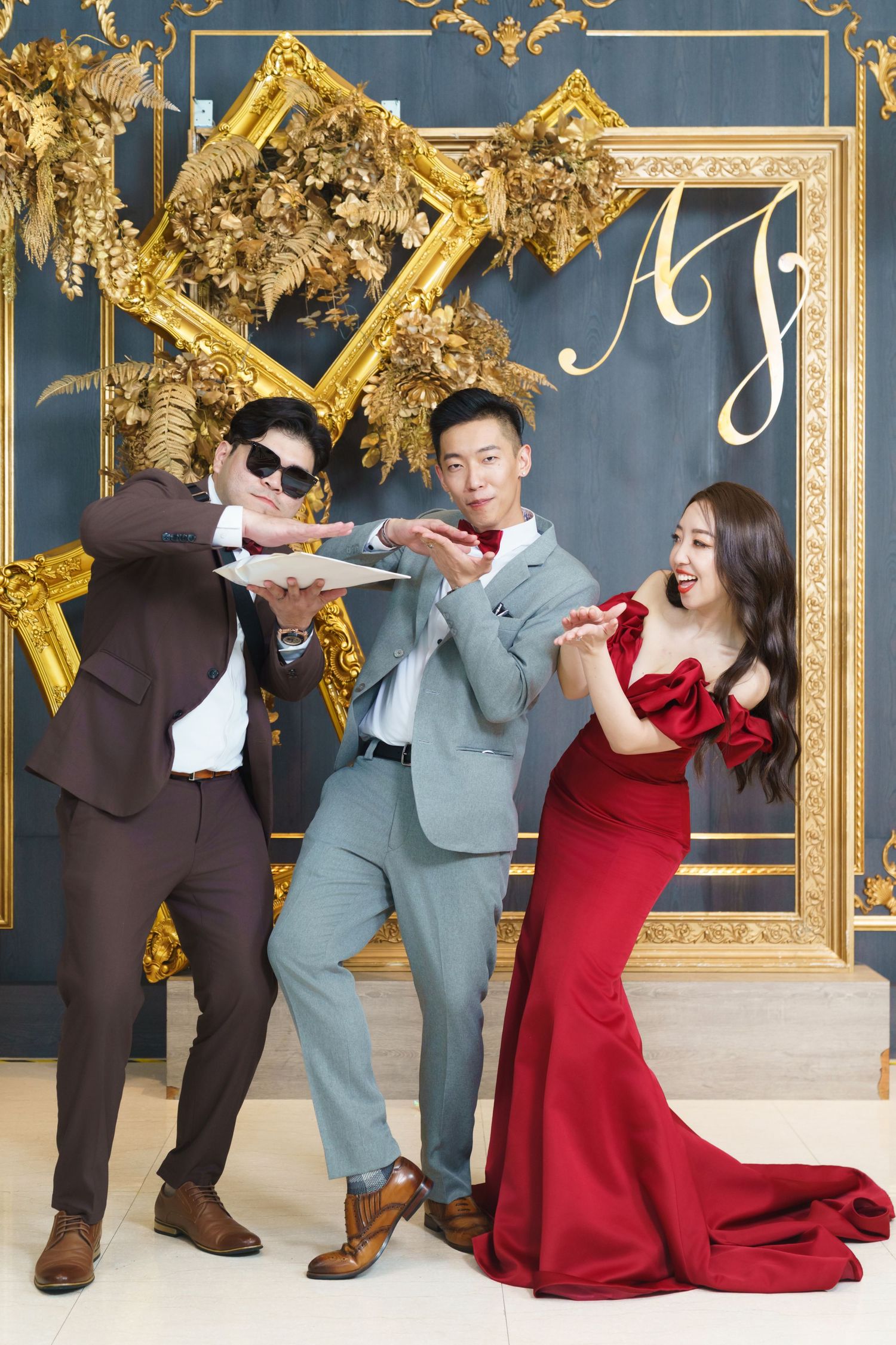 Three people posing playfully against an elegant backdrop with gold frames, wearing formal attire in brown, gray and red.