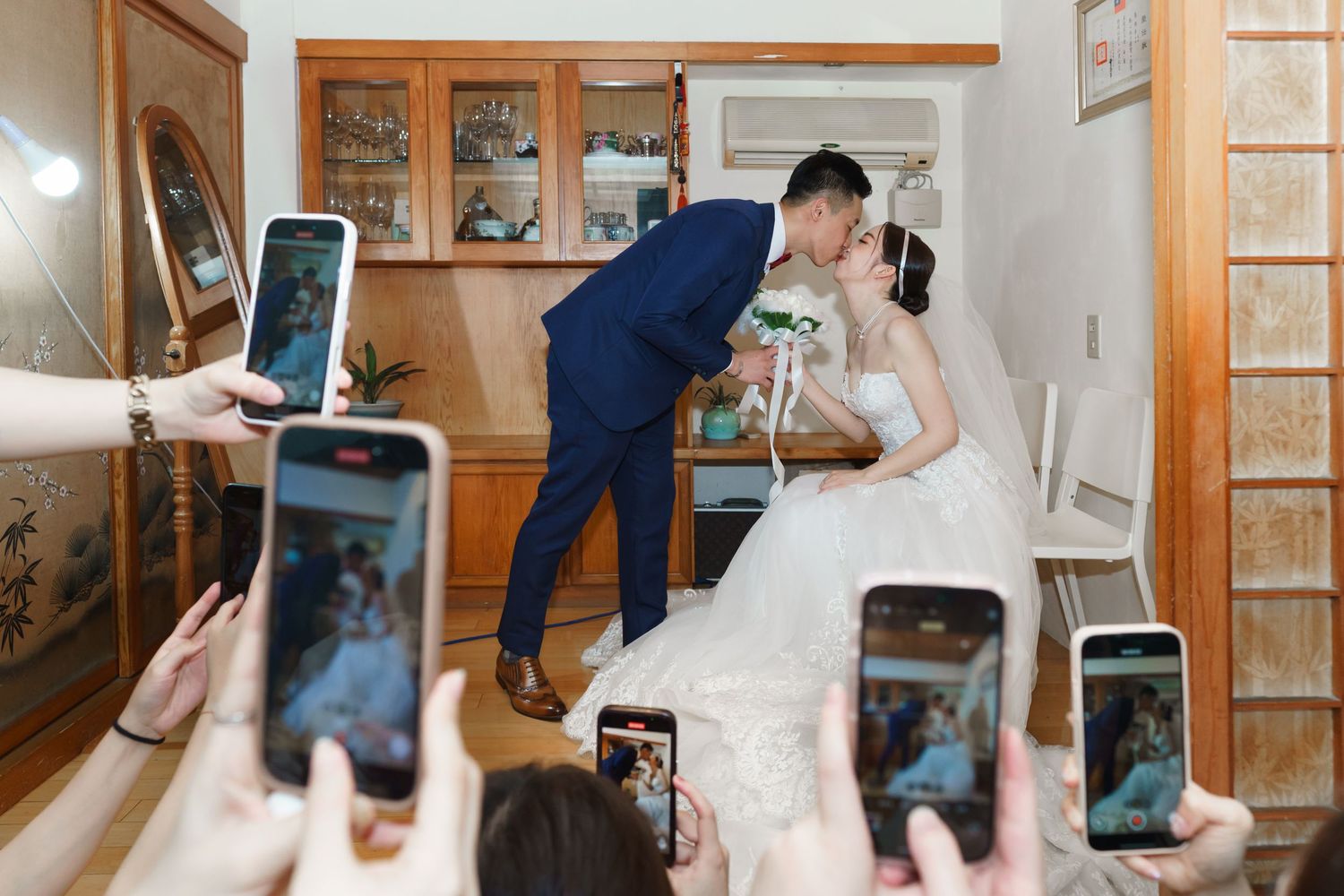 Multiple phones capture a special moment between a couple in formal wedding attire at an indoor venue.
