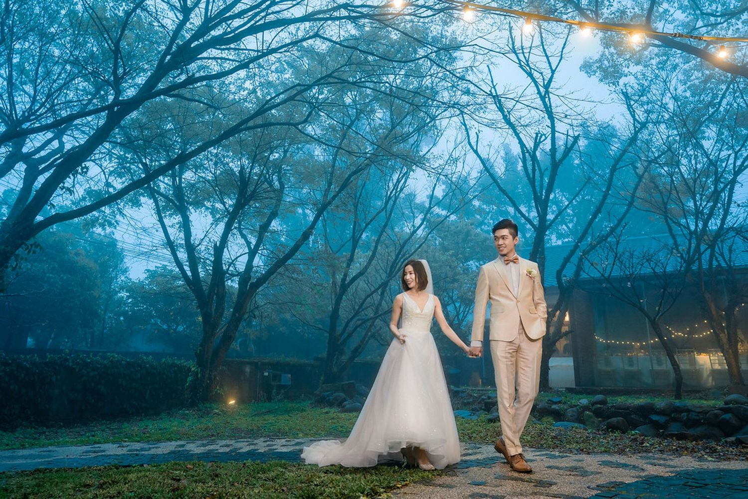 A couple walks hand in hand along a garden path at dusk with glowing lights and misty blue trees creating a dreamy atmosphere.