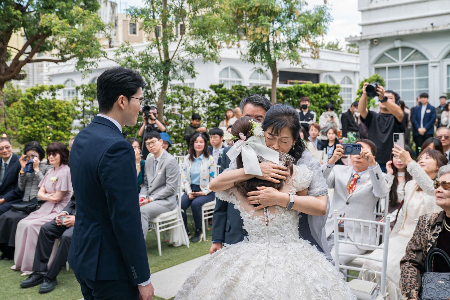 Guests gather outdoors at an elegant garden wedding ceremony with white chairs and lush greenery in the background.