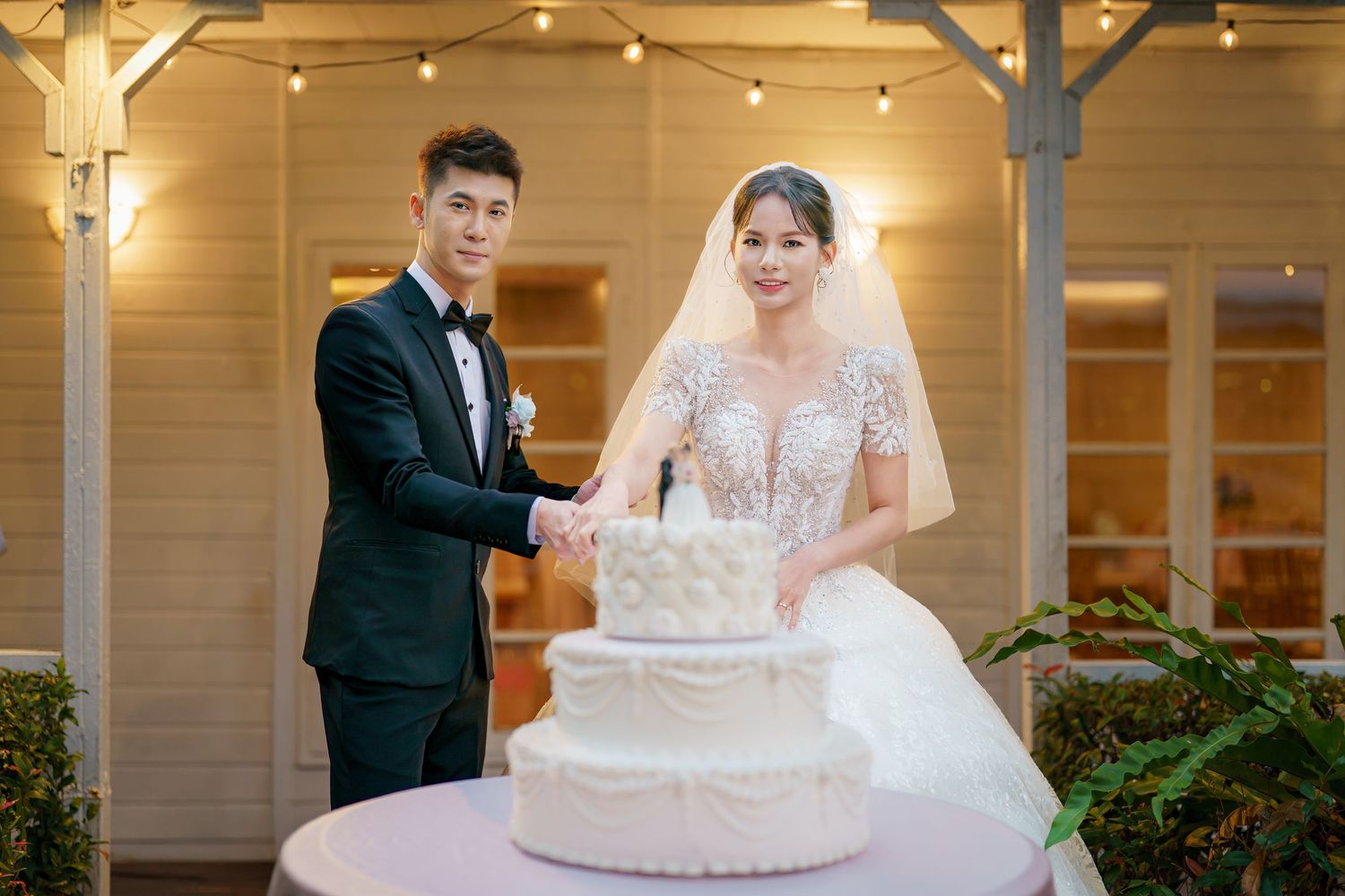 A wedding couple cuts their elegant white tiered cake together at an indoor reception venue.