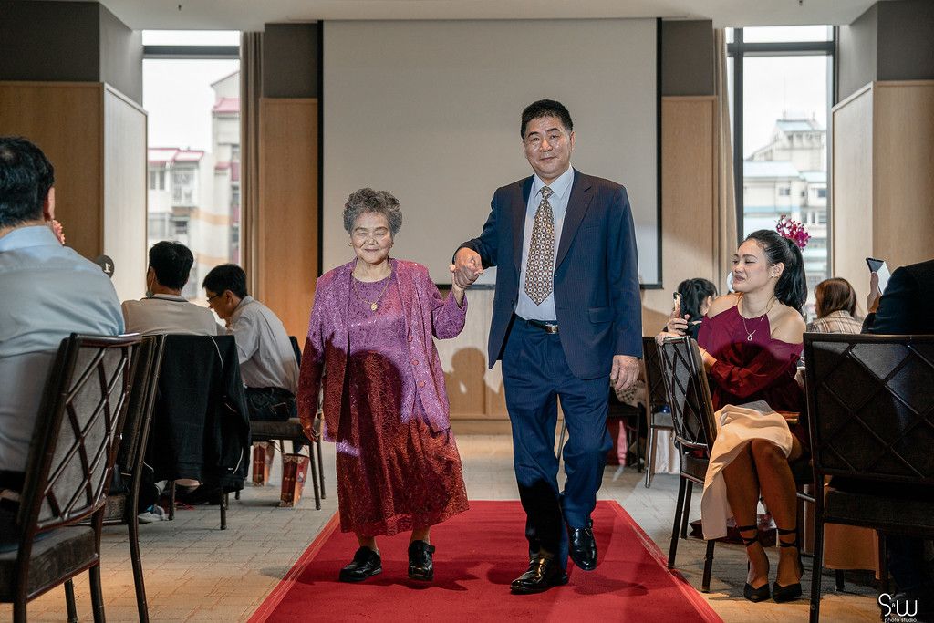 A pair walks arm in arm down a red carpet aisle during a wedding reception.