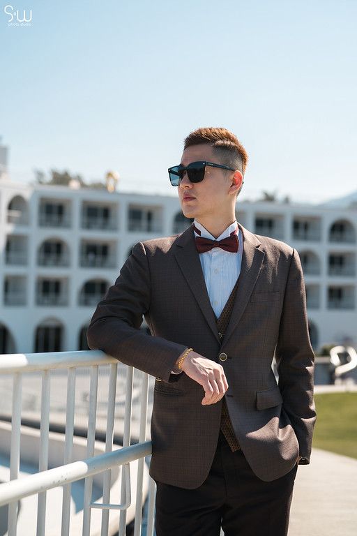 Stylish groom in sunglasses and brown suit stands against white hotel facade.