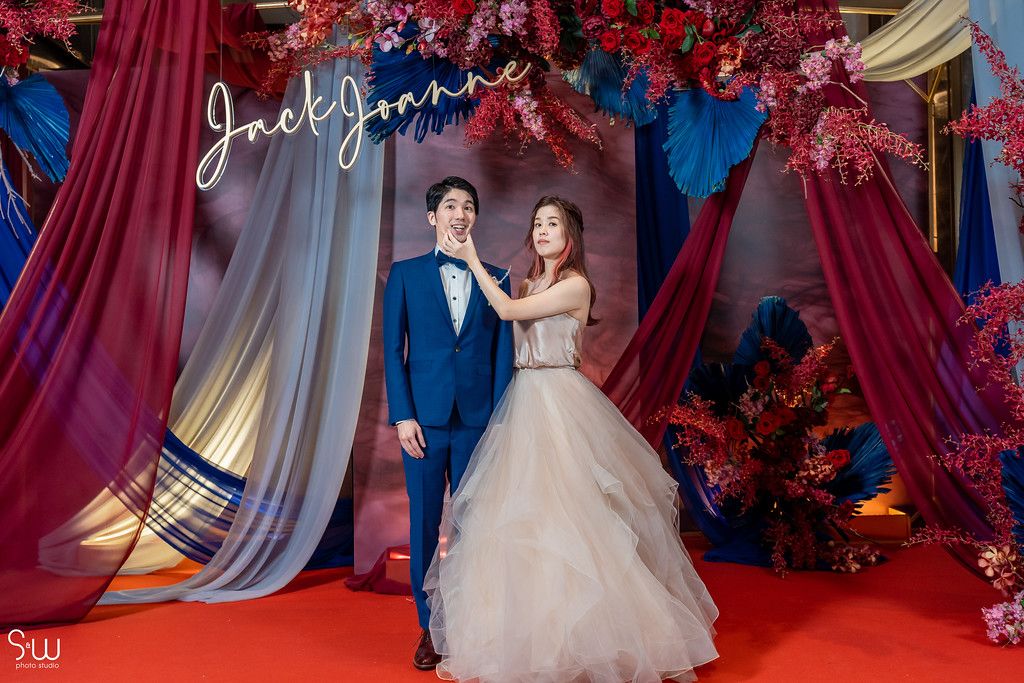 Elegant posed wedding portrait against dramatic backdrop with red florals and flowing drapes.