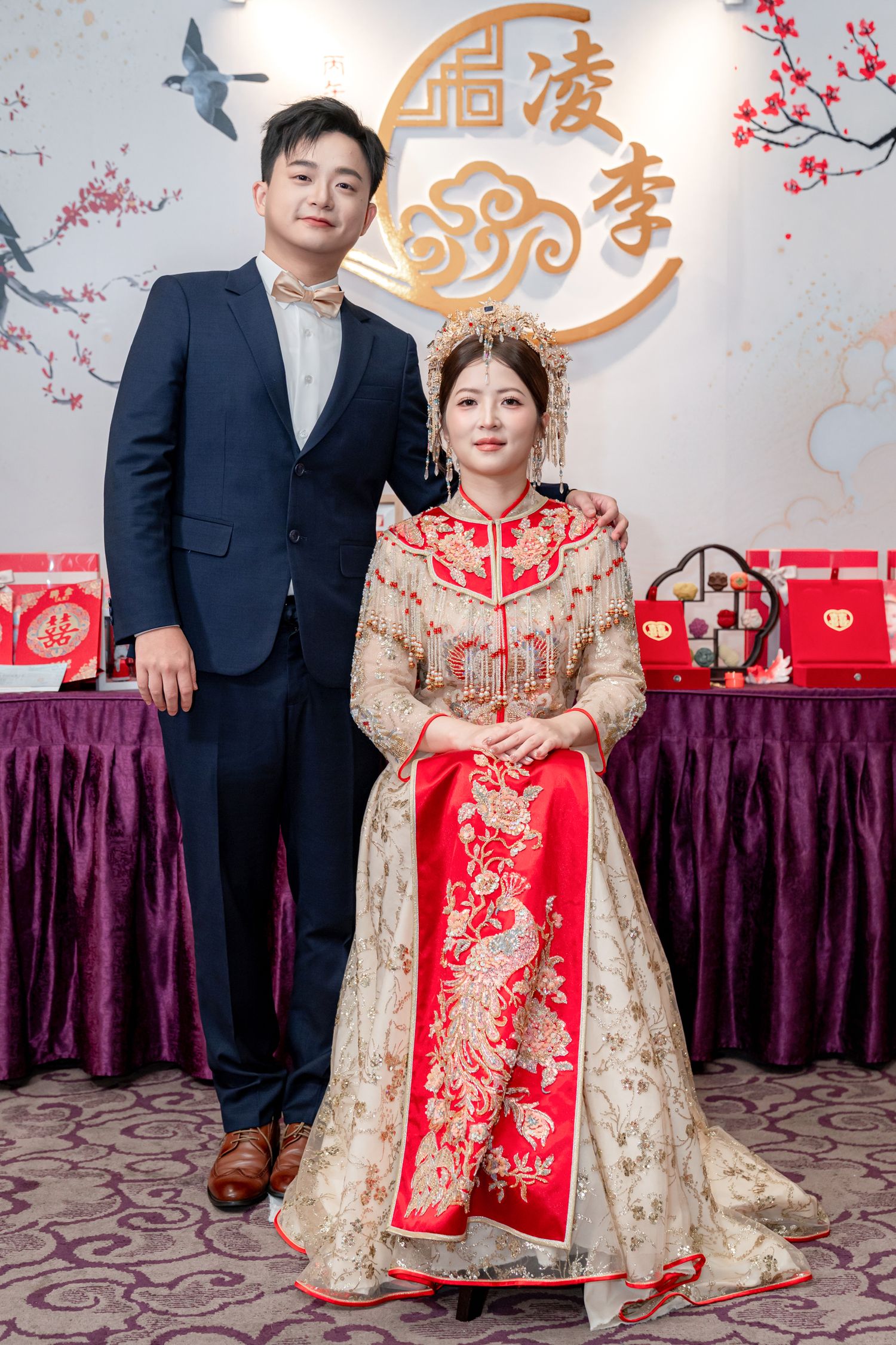 Bride in a red and gold embroidered Chinese wedding gown with phoenix headdress sits beside her groom in a navy suit at a tea ceremony.