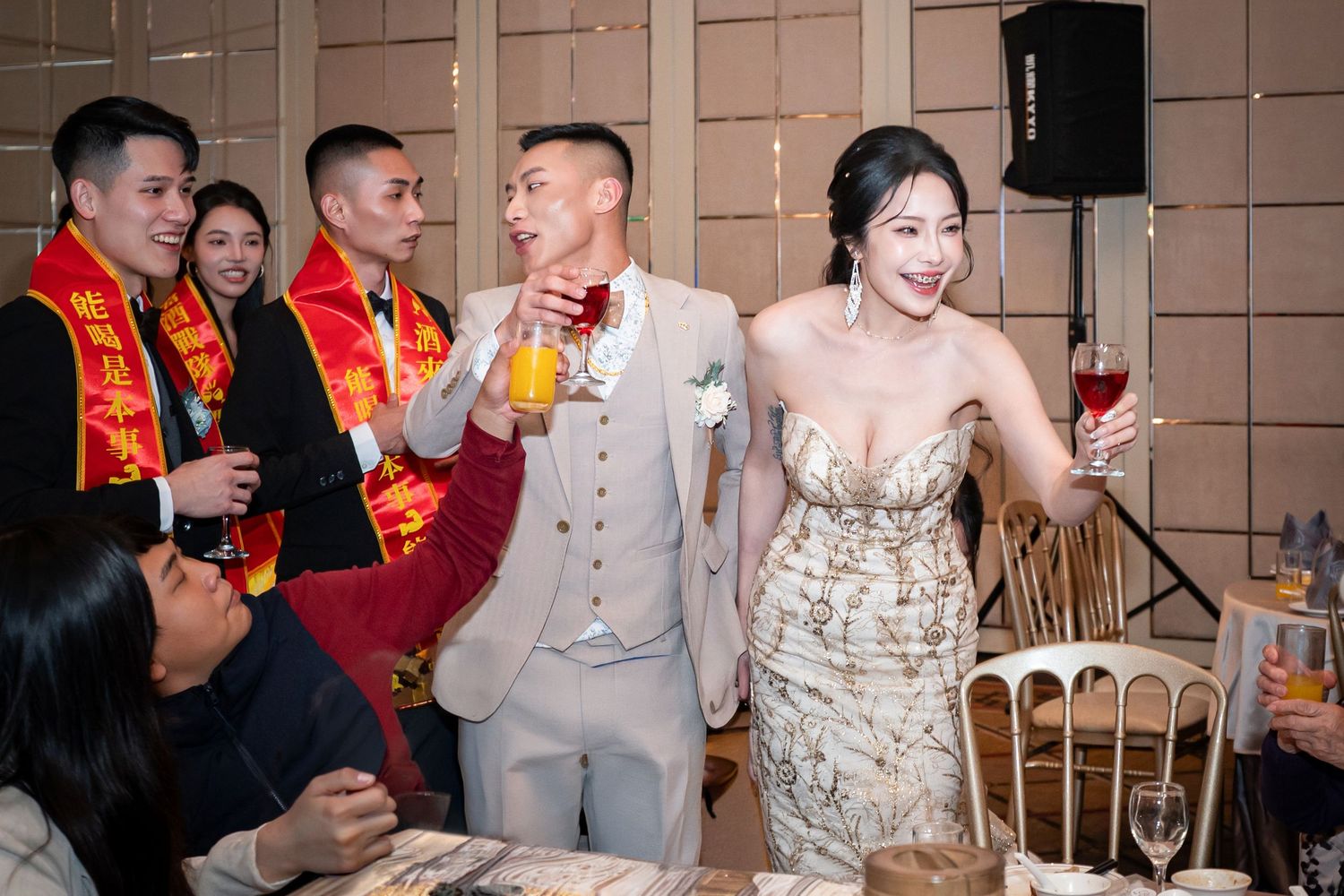 Wedding celebration with people toasting drinks at a reception while wearing traditional red sashes and formal attire.