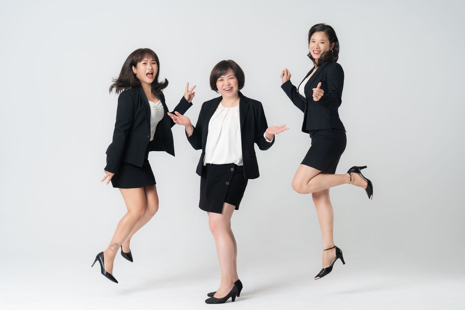 A group of women in black blazers and heels pose playfully in a bright studio setting.