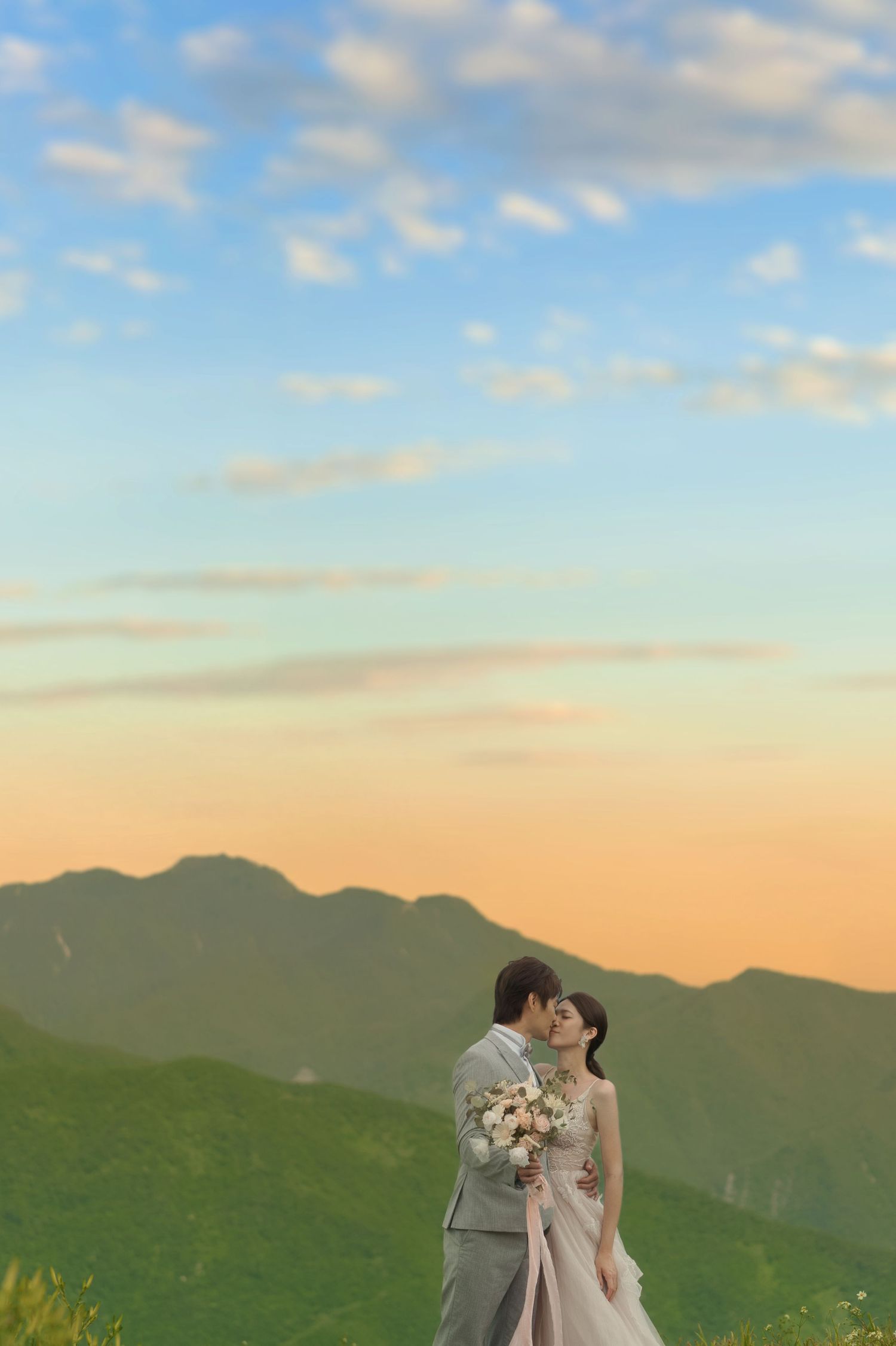 Couple shares romantic moment against backdrop of green mountains and sunset sky during outdoor wedding photoshoot.