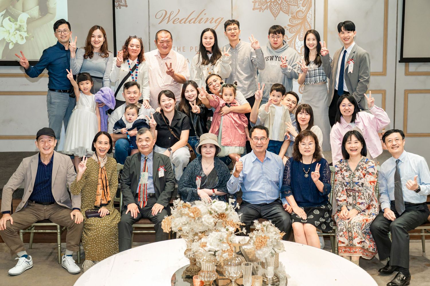 Wedding guests gather for group photos in an elegant venue with floral decorations and beige tones.
