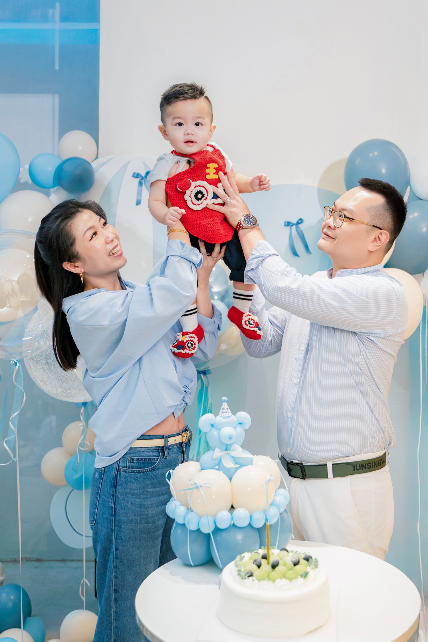 A joyful family celebrates a first birthday party with blue balloons and decorations in a bright studio setting.