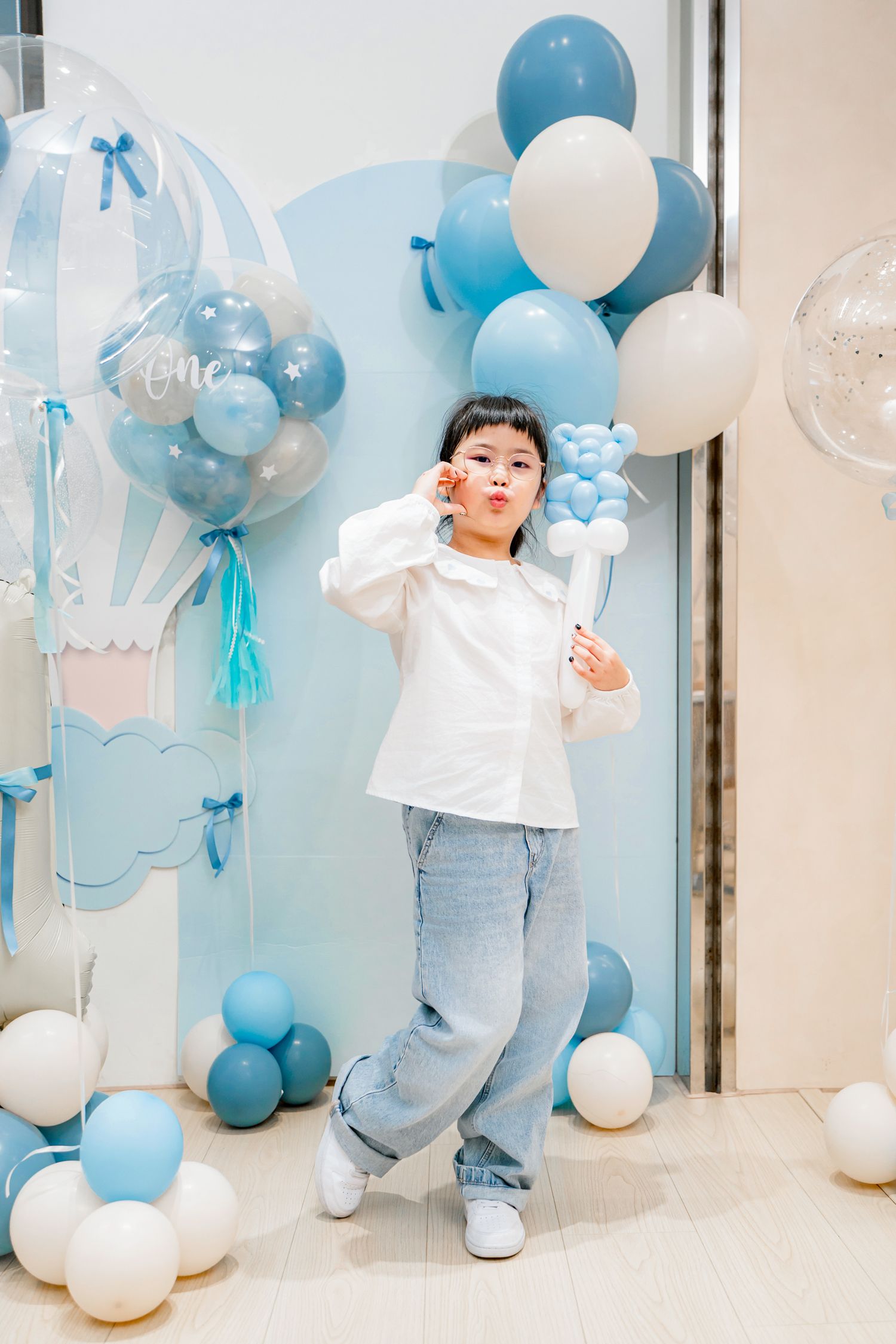 A baby in white and blue outfit plays among blue and white balloon decorations at a celebration party.