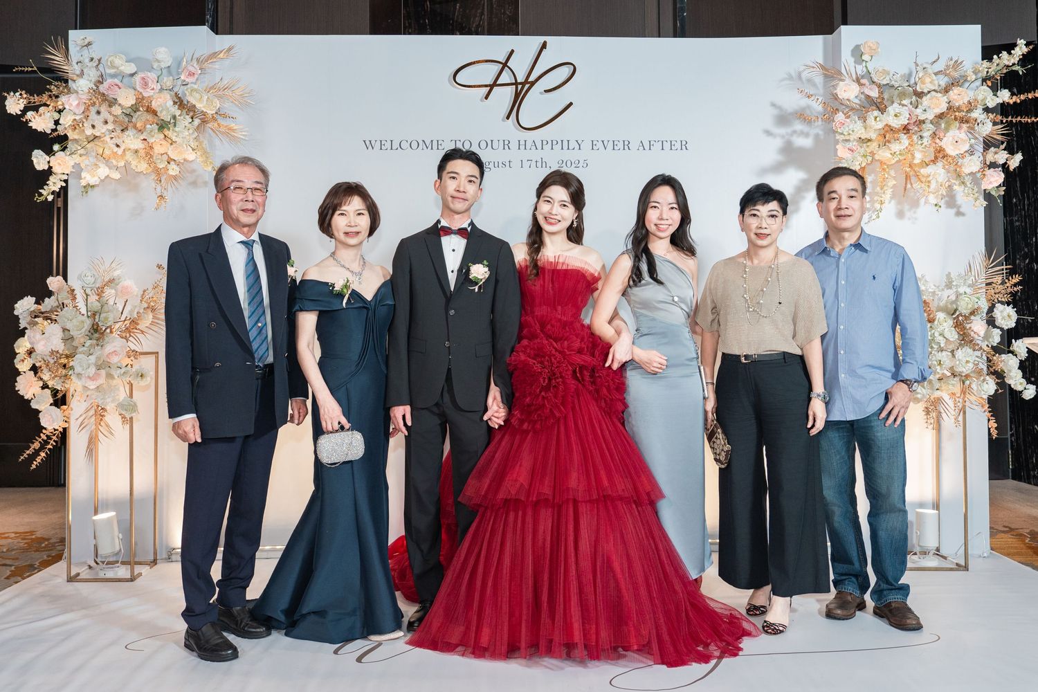 Elegant wedding party posing in formal attire against a floral backdrop with red gown as centerpiece.