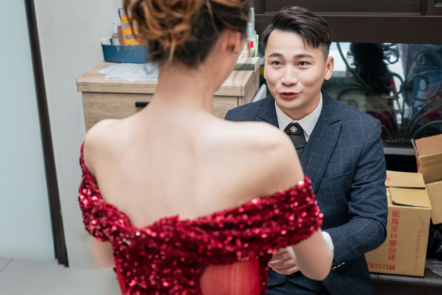 A couple shares a moment as the woman wears a red sequined off-shoulder gown and the man wears a navy suit.