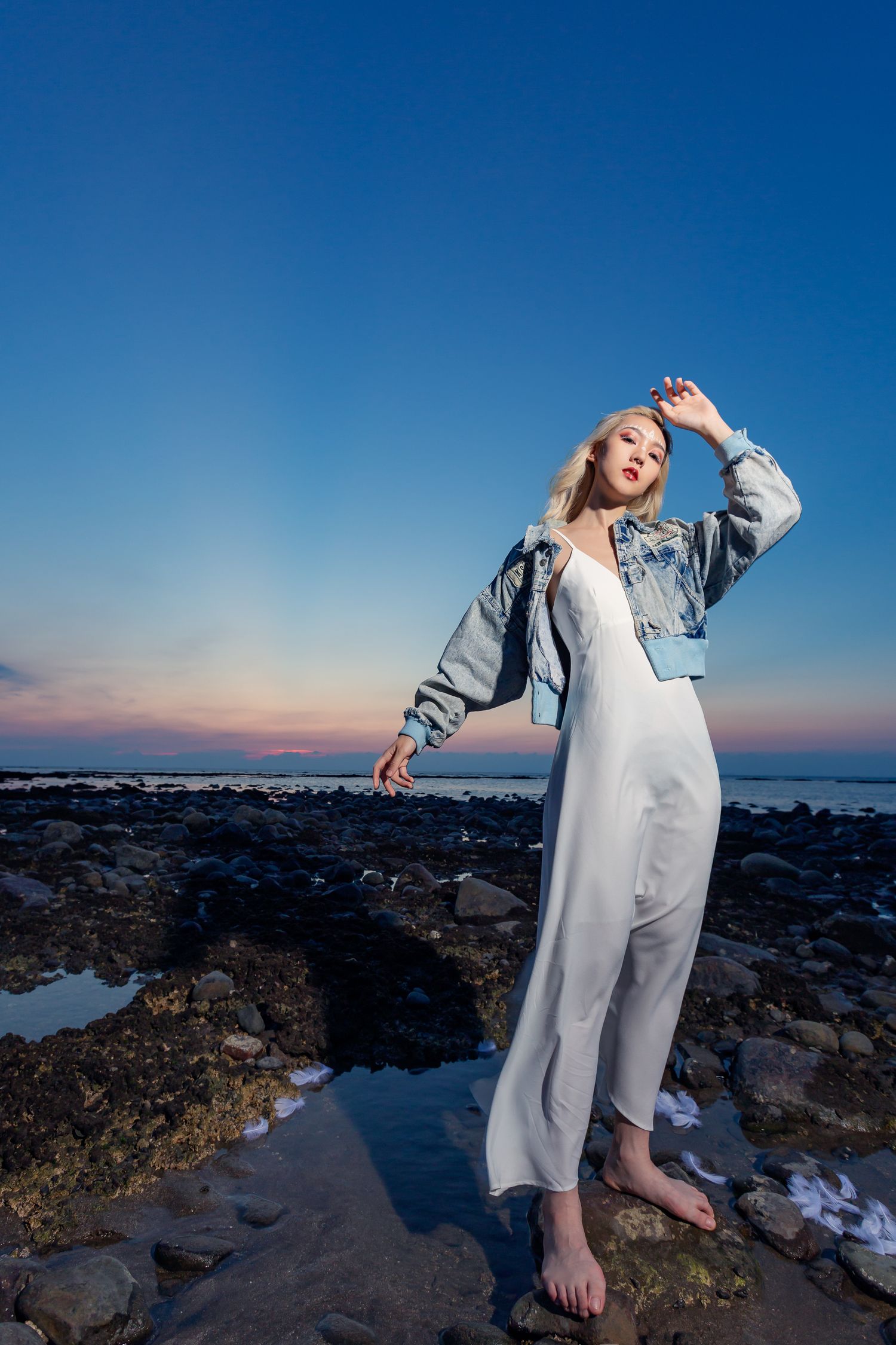 Person in white dress and denim jacket poses barefoot on rocky coastal shore at sunset against blue sky.