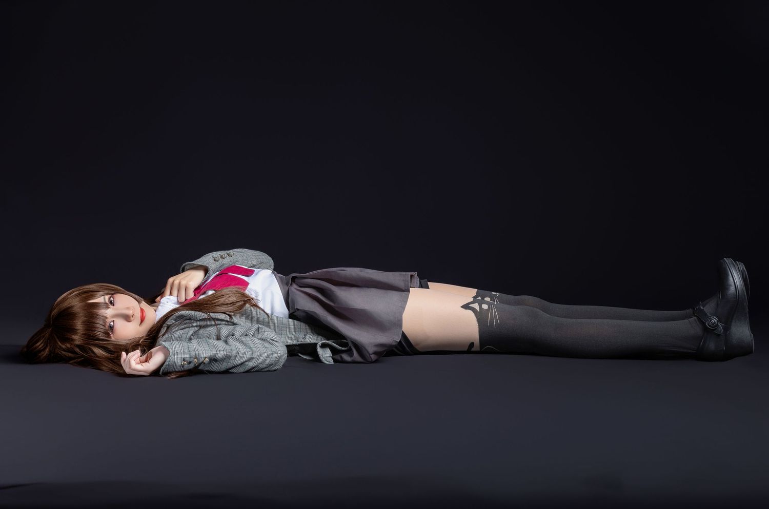 Someone in gray attire lies down with arms crossed on a dark floor with dramatic studio lighting.