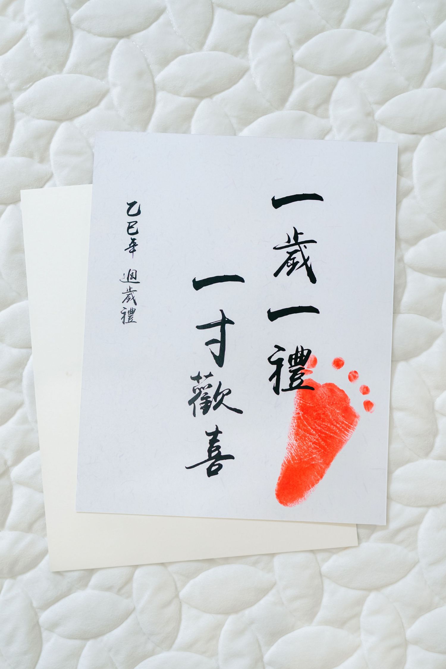 Japanese calligraphy artwork featuring black characters and a bright red baby footprint on white paper against textured background.