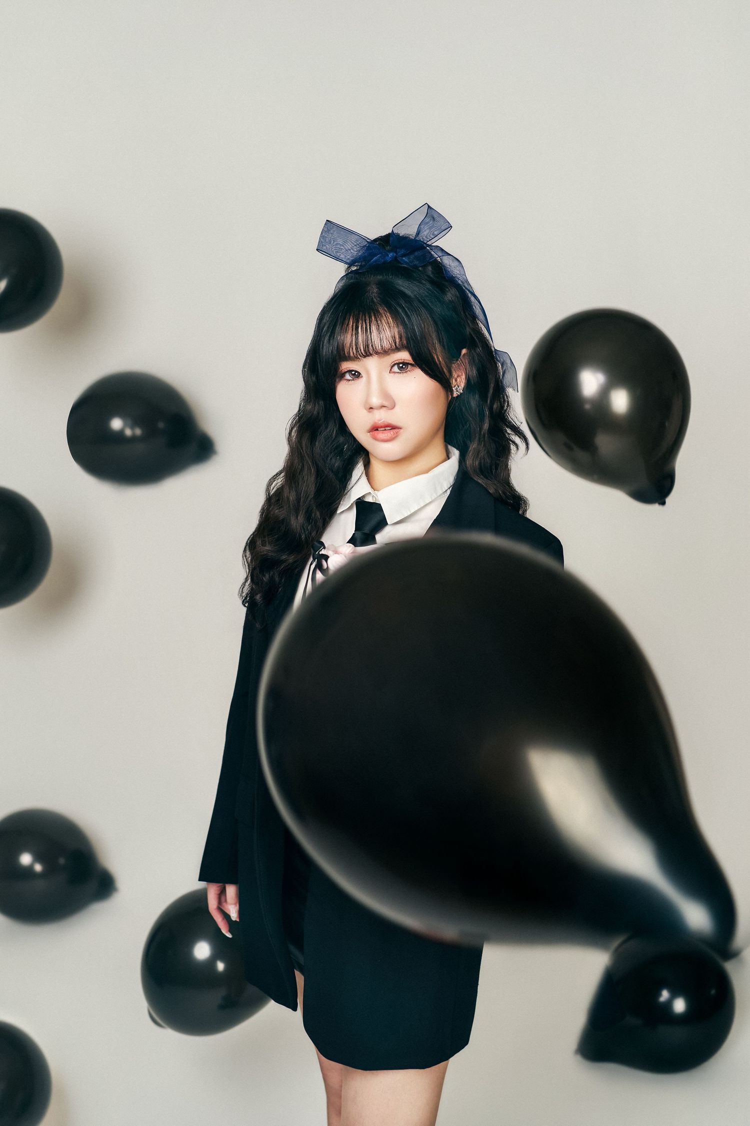 Young woman in black blazer and skirt poses with black balloons in a studio, wearing a navy bow hair accessory.