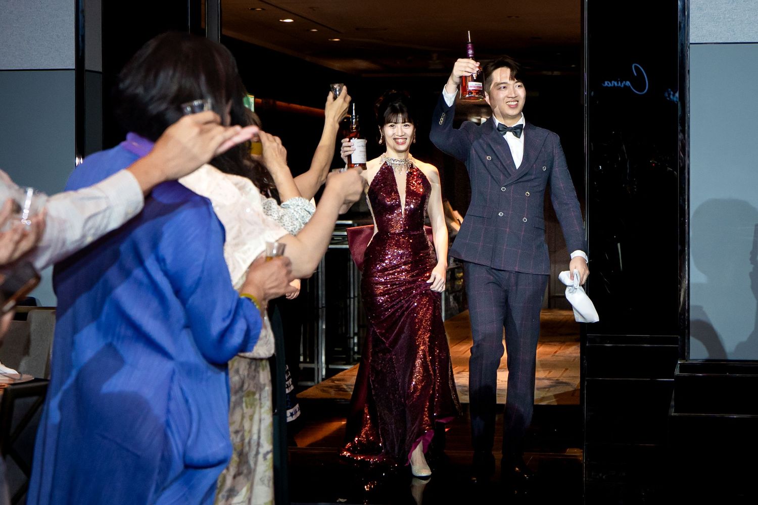 Elegant couple in formal attire entering a celebration, woman in red gown, man in blue suit, guests raising glasses.