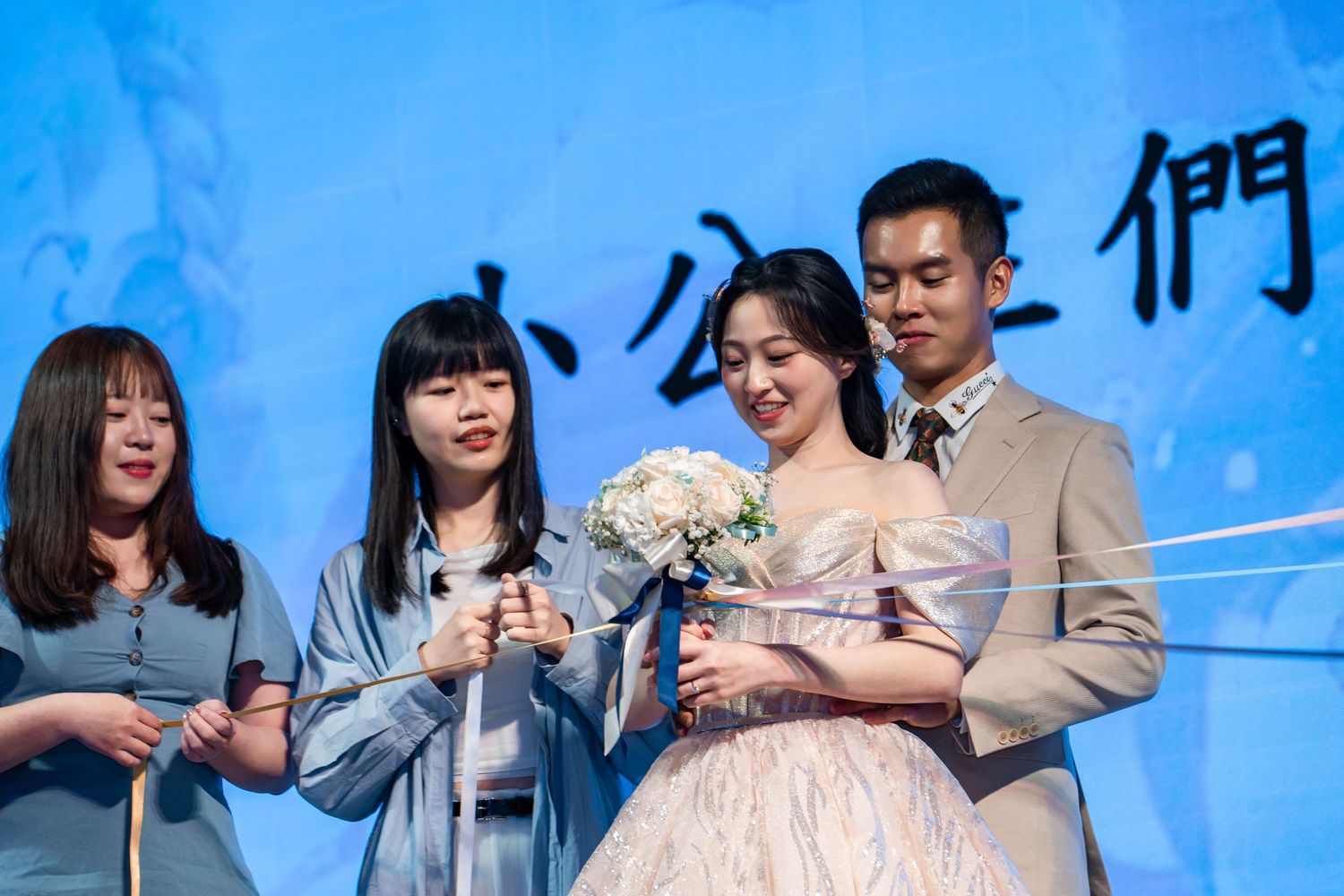 Bride holding bouquet with groom and bridesmaids at Asian wedding ceremony on stage with blue backdrop.