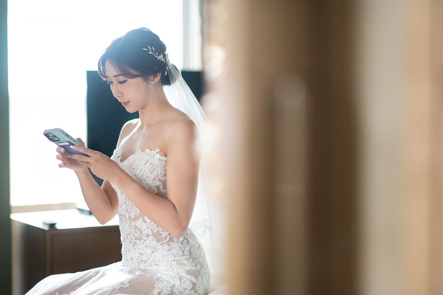 A bride in a strapless lace wedding gown checks her phone while sitting near a window in soft natural light.