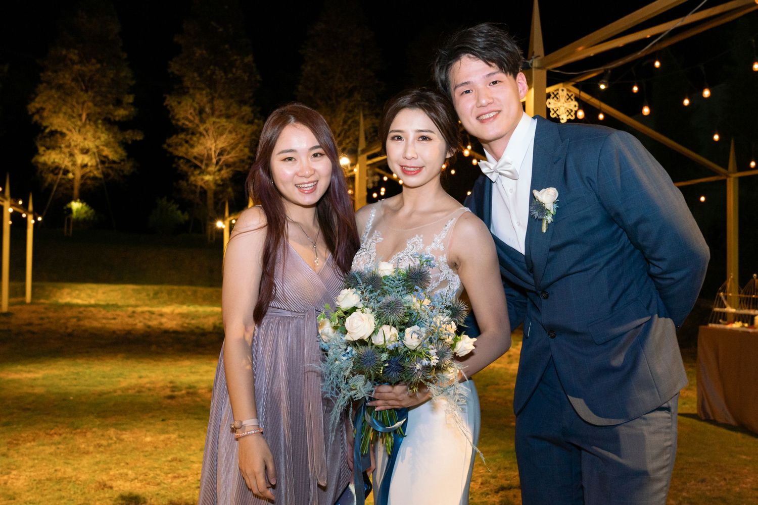 Three wedding guests pose together outdoors at night under string lights with trees in the background.