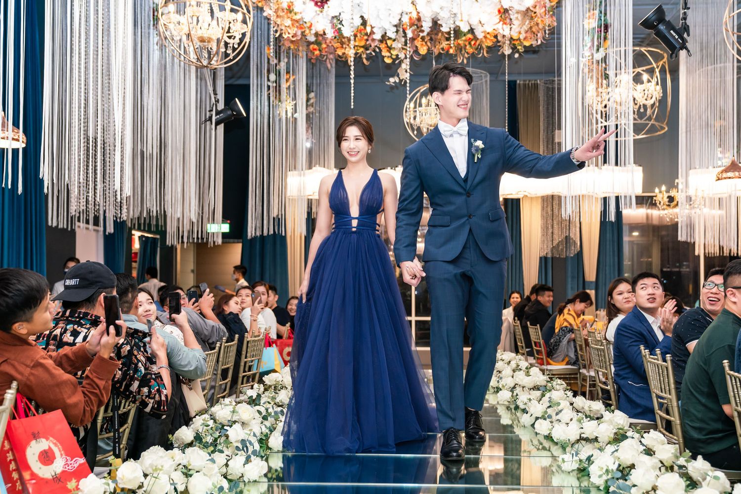 Couple celebrates on a stage in a glamorous ballroom with crystal chandeliers and floral decorations.