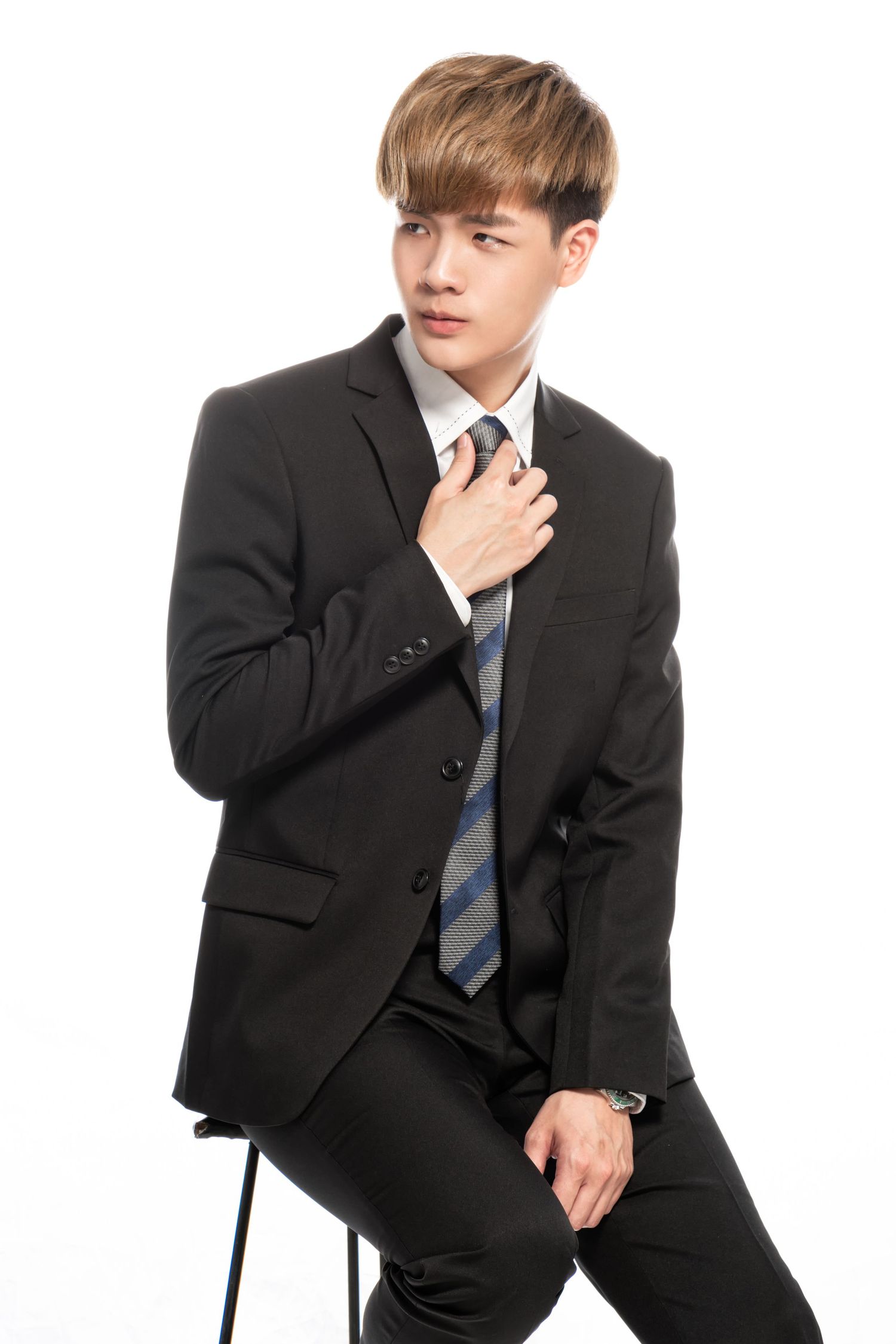 A young professional in a dark suit adjusts his striped tie while sitting on a stool against a white background.