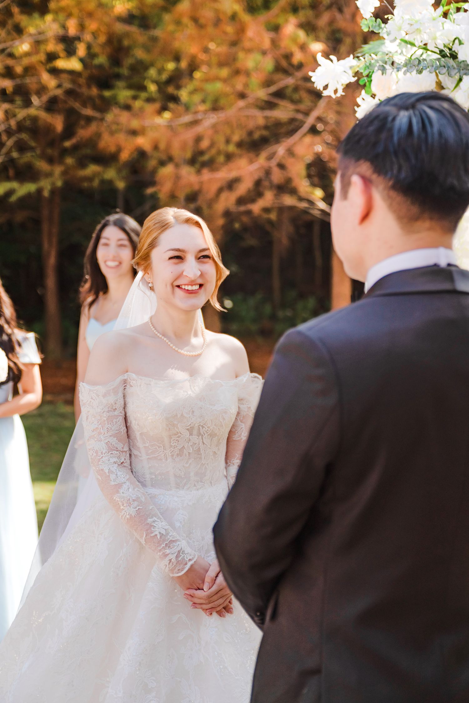 An outdoor autumn wedding ceremony with a bride in a long-sleeved lace gown exchanging vows on a sunny day.