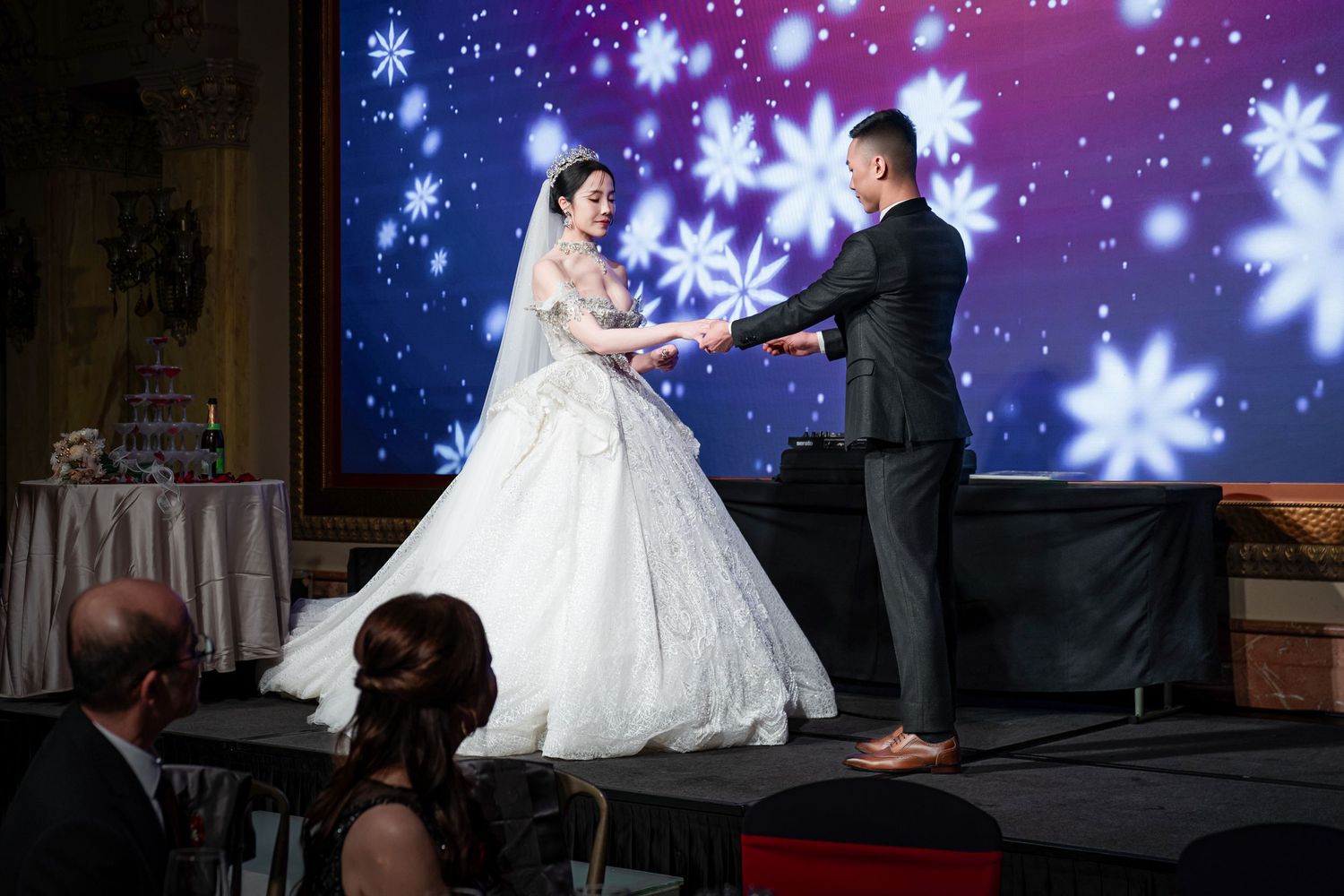 A couple performs their first dance on a stage with illuminated snowflake projections in the background.