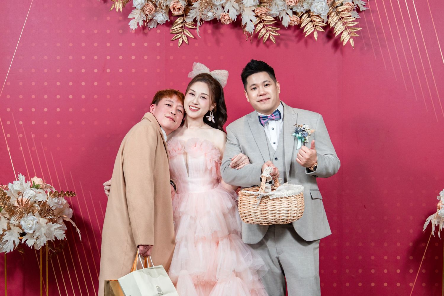 Three people in formal wear share a joyful moment against a pink backdrop with floral decorations above.