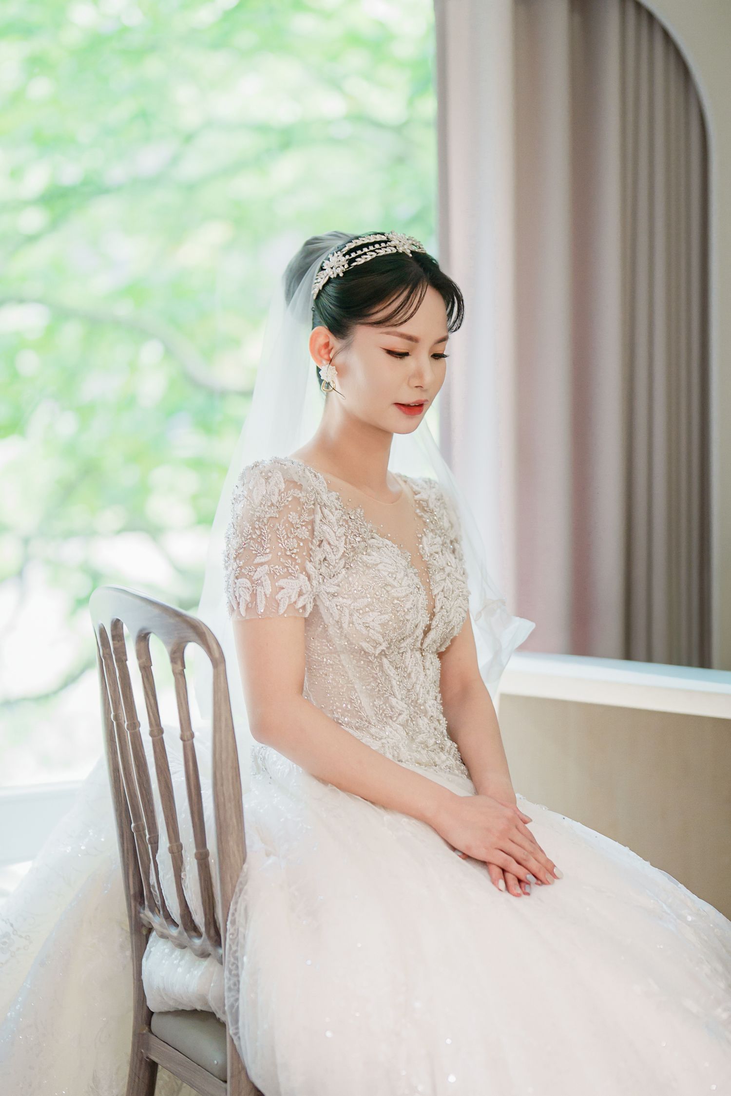 A bride in an elegant lace wedding dress sits gracefully near a window with soft natural lighting.