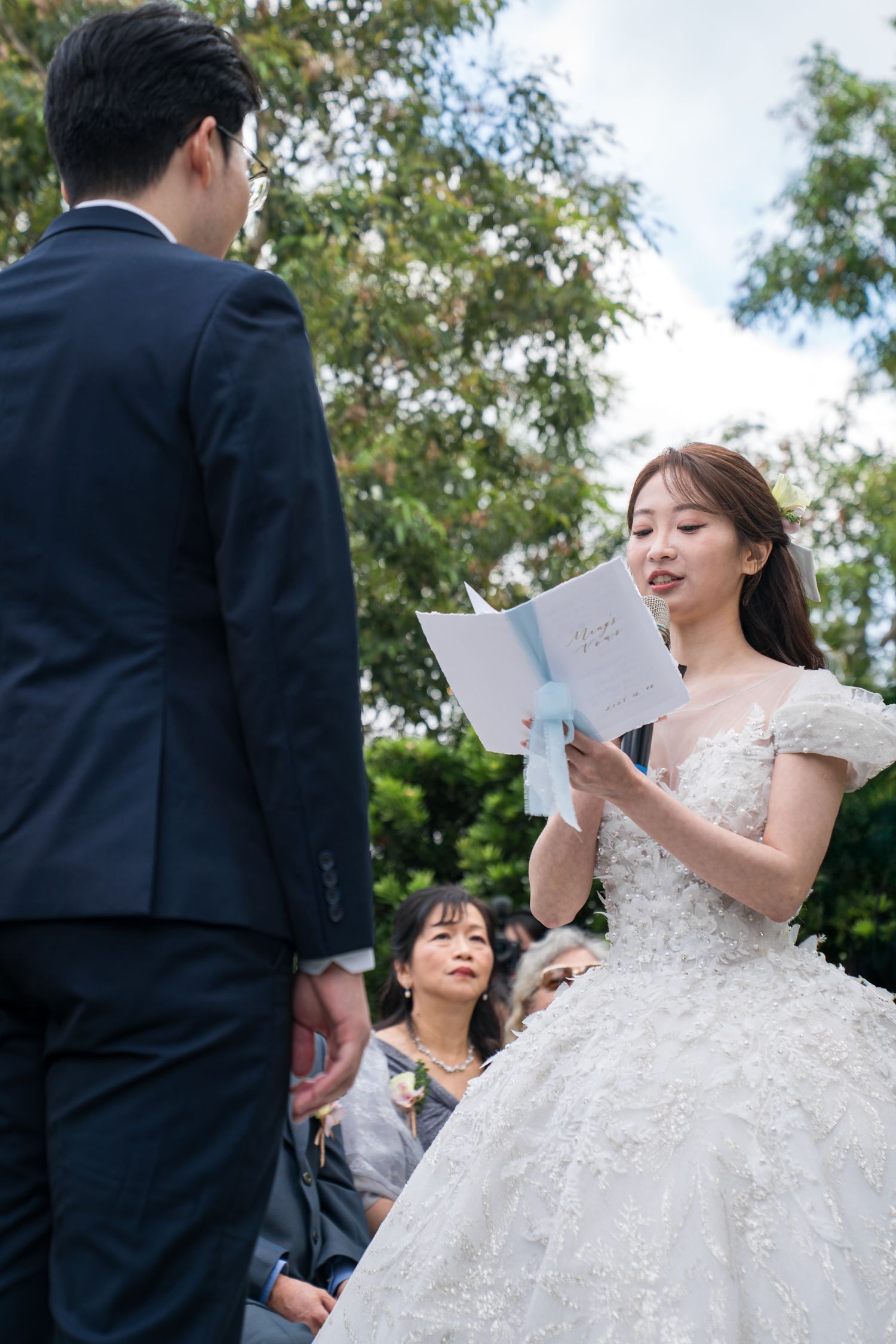 A bride in a white lace gown reads from a paper during an outdoor wedding ceremony with trees in the background.