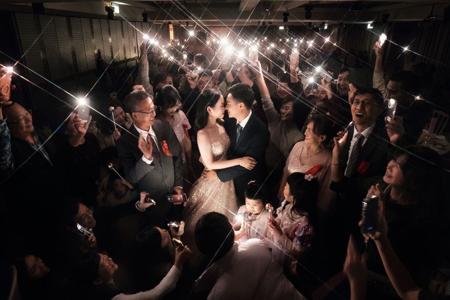 Crowd of concert attendees in a dark venue illuminated by flashing lights and phone screens during a live performance.