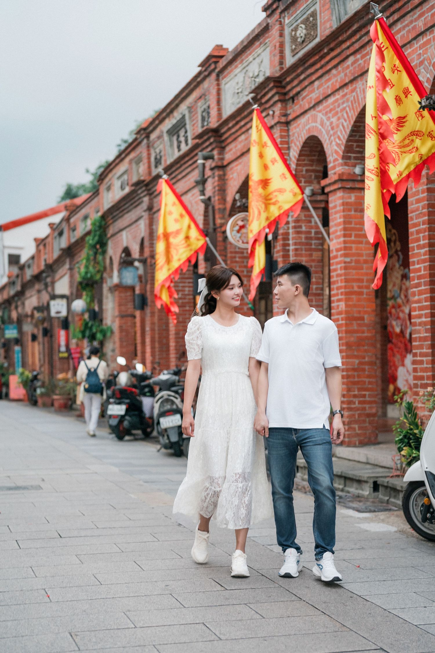 Two people in white outfits walk down a historic brick street lined with red flags and traditional architecture.