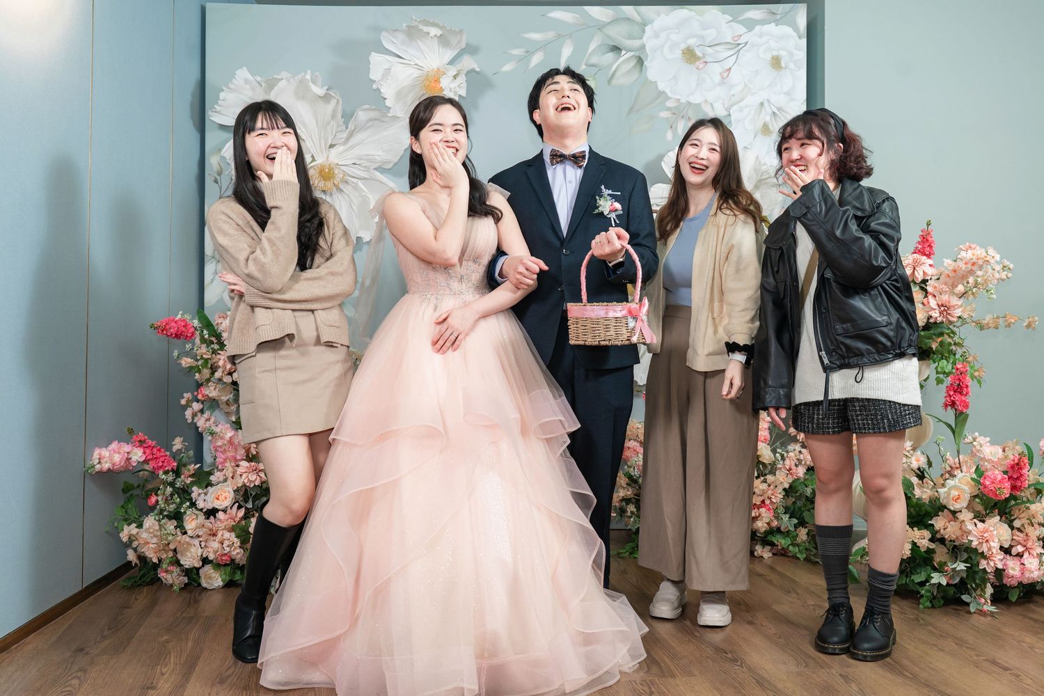 A group of people pose together in formal attire with pink floral decorations in the background.