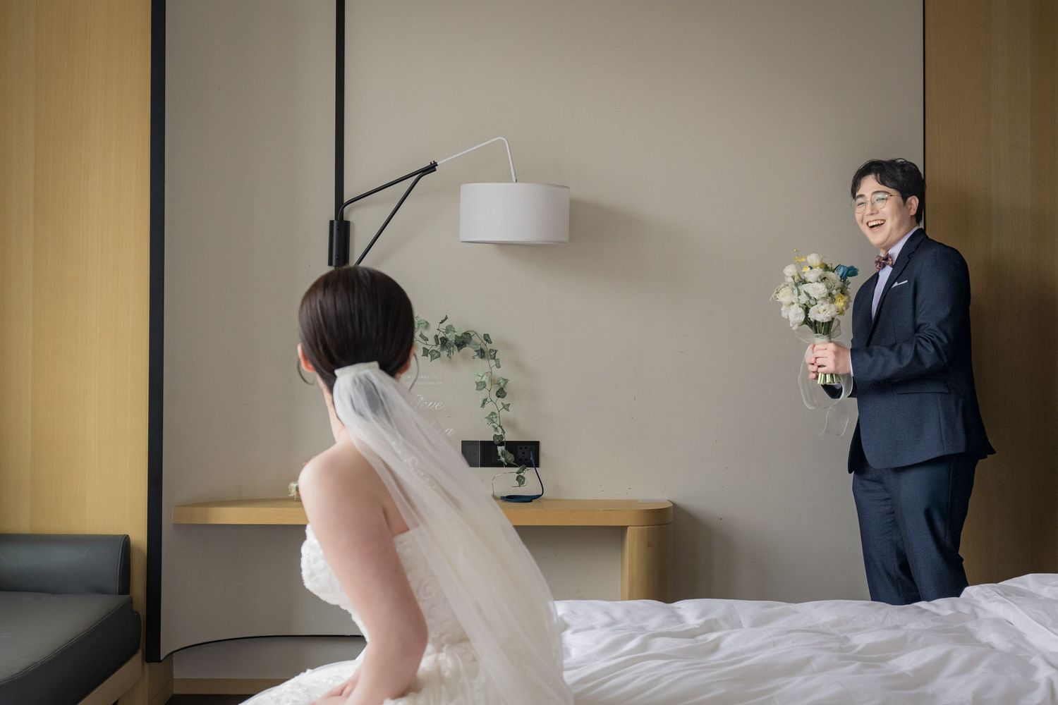 A bride in a white wedding dress and veil stands in a minimalist room with beige walls and modern decor.