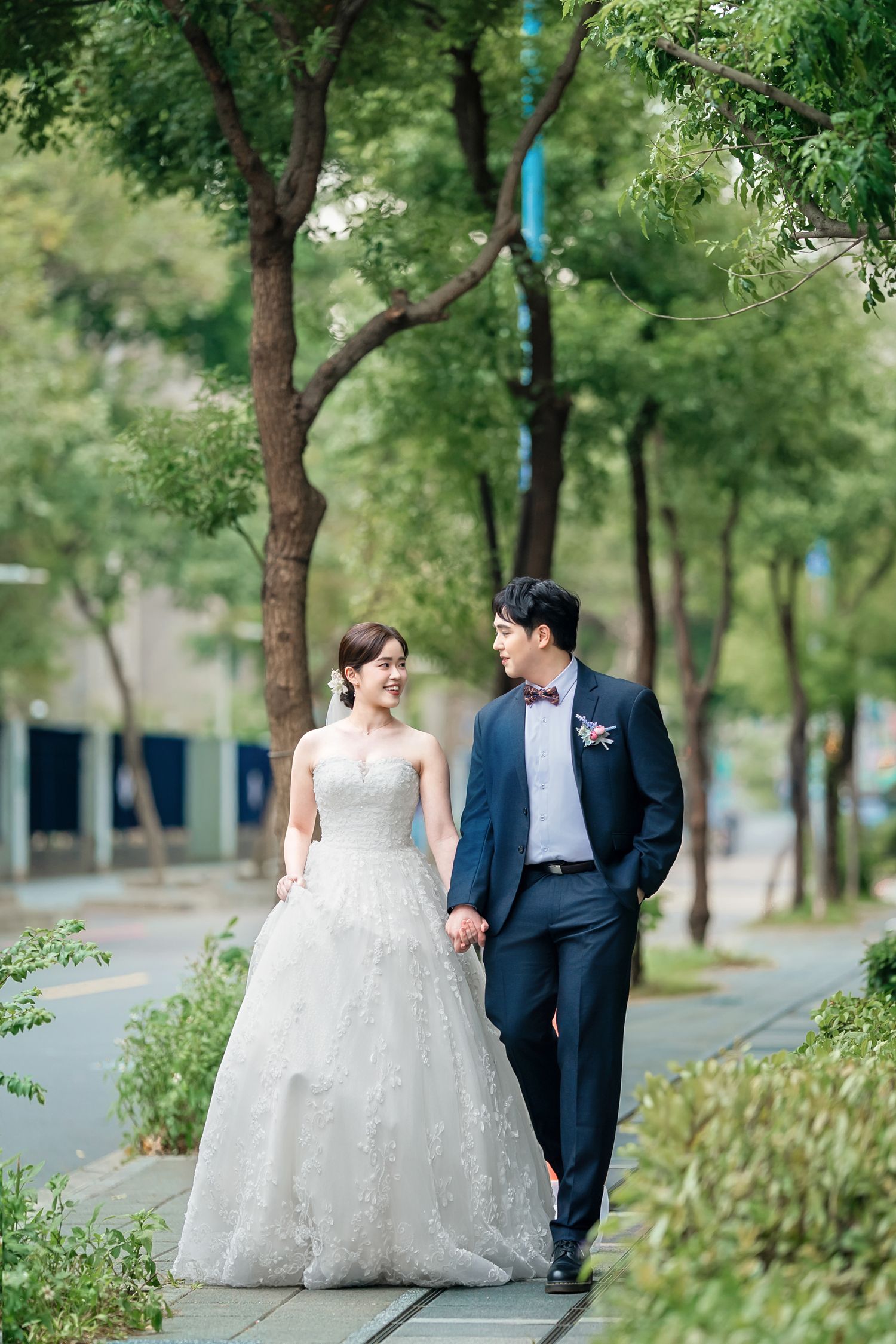 A couple in wedding attire walks together on a tree-lined path surrounded by lush greenery on a sunny day.
