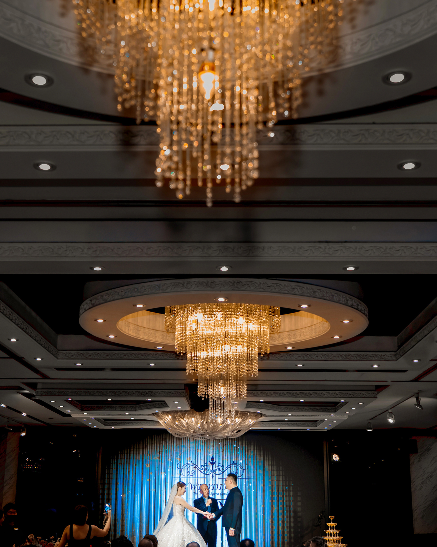 Elegant wedding ceremony under ornate crystal chandeliers with a blue backdrop in a luxurious ballroom setting.