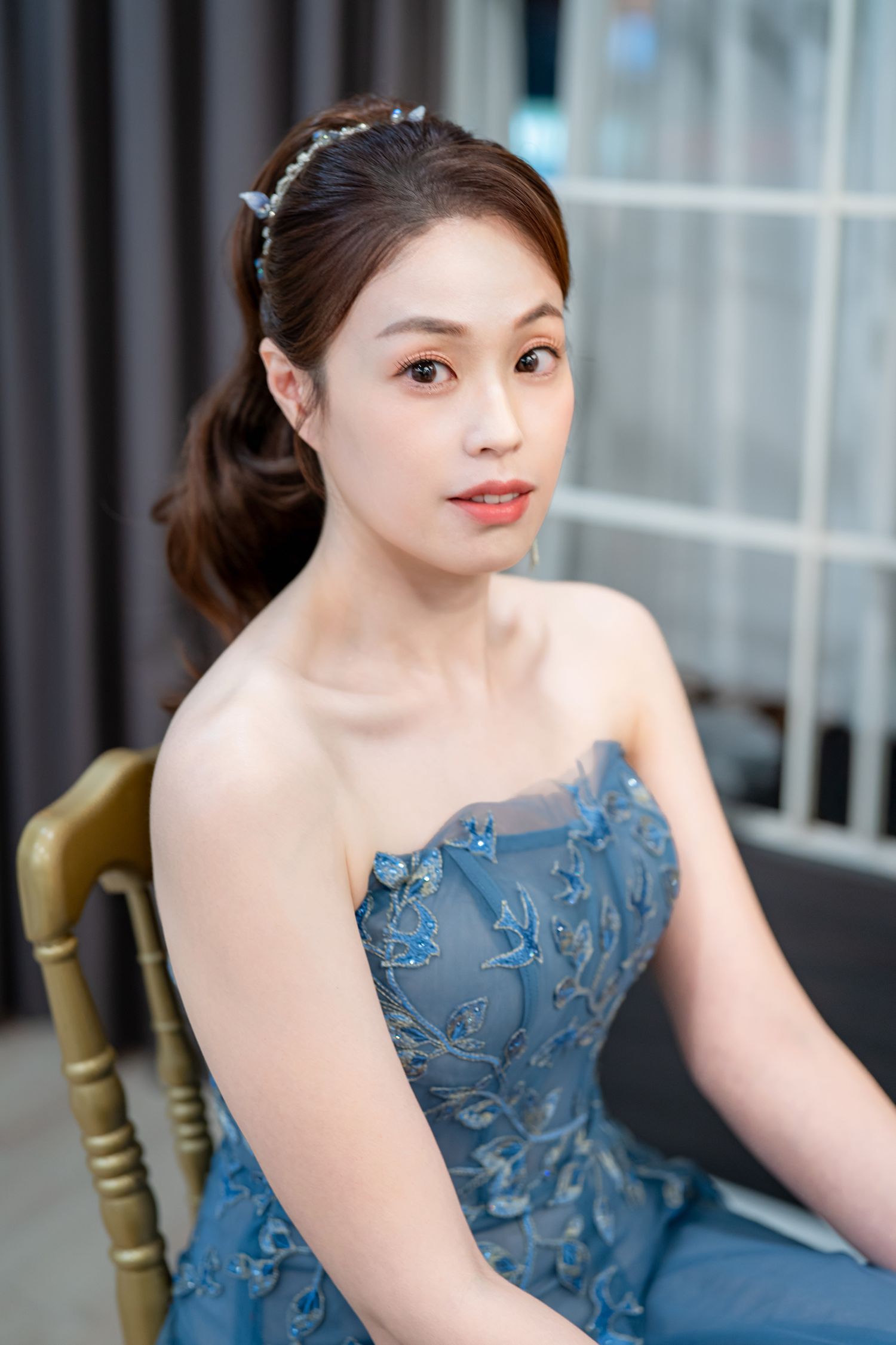 A person in an elegant blue lace evening gown poses gracefully on a chair in an indoor setting.