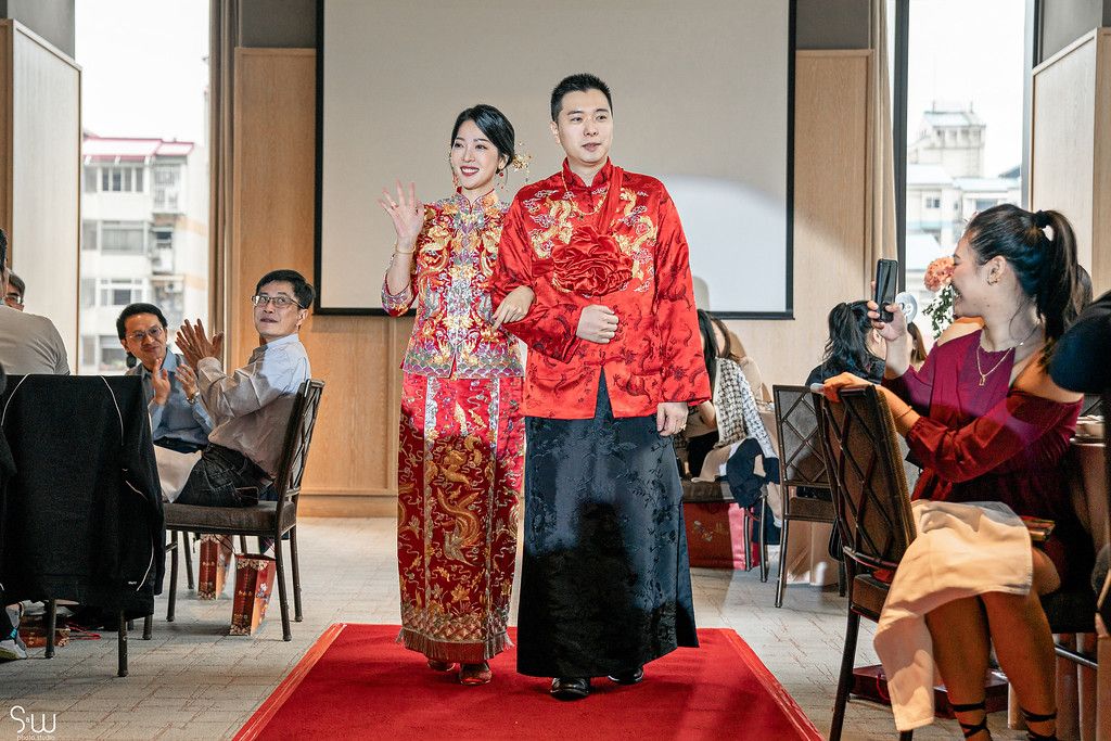 A wedding ceremony shows a couple in red traditional attire walking down a red carpet aisle.