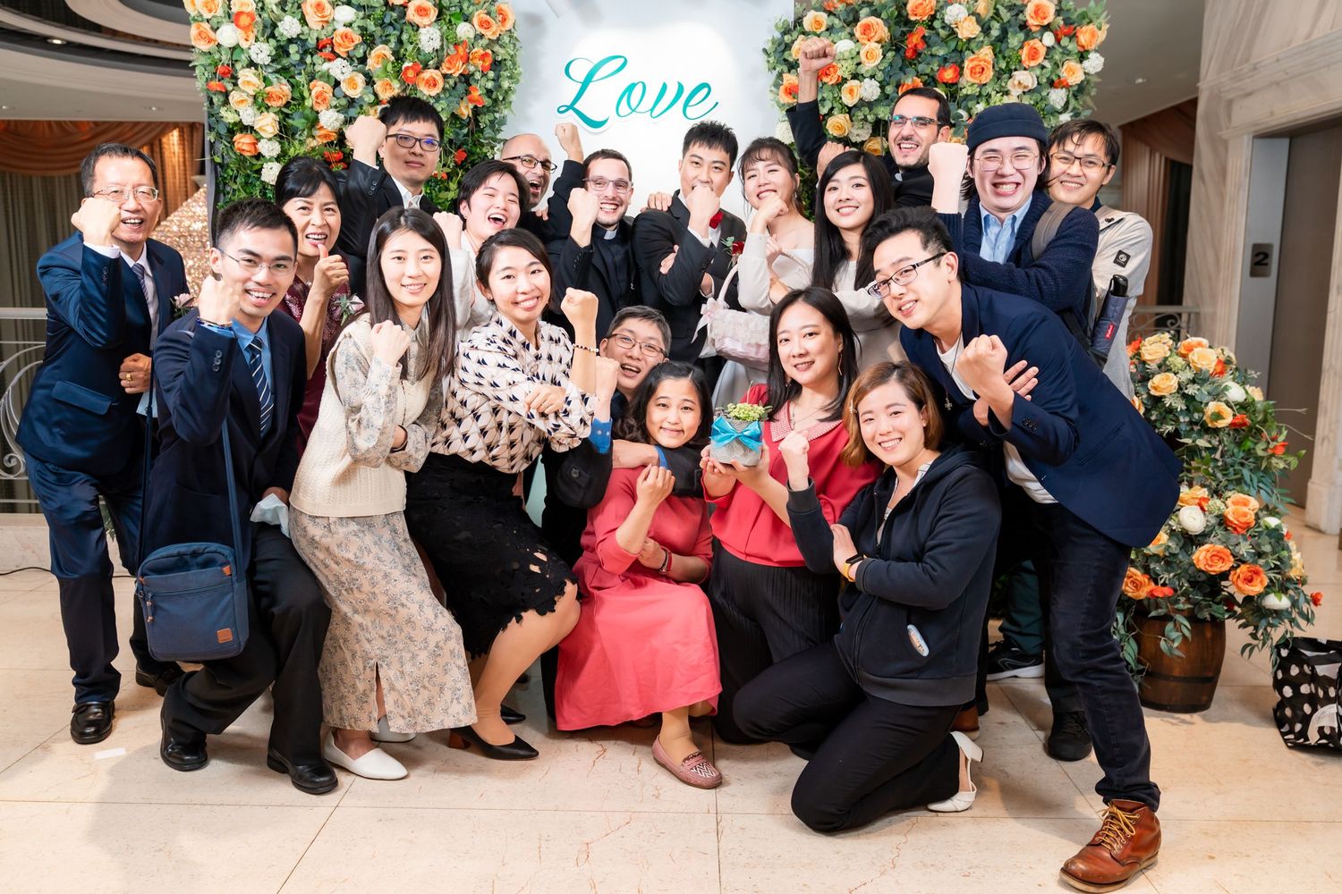 Large group photo with people in business attire posing casually in front of floral decorations.
