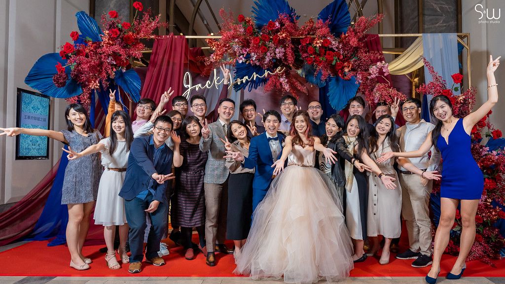 Wedding party group photo with celebratory poses against red and blue floral backdrop.