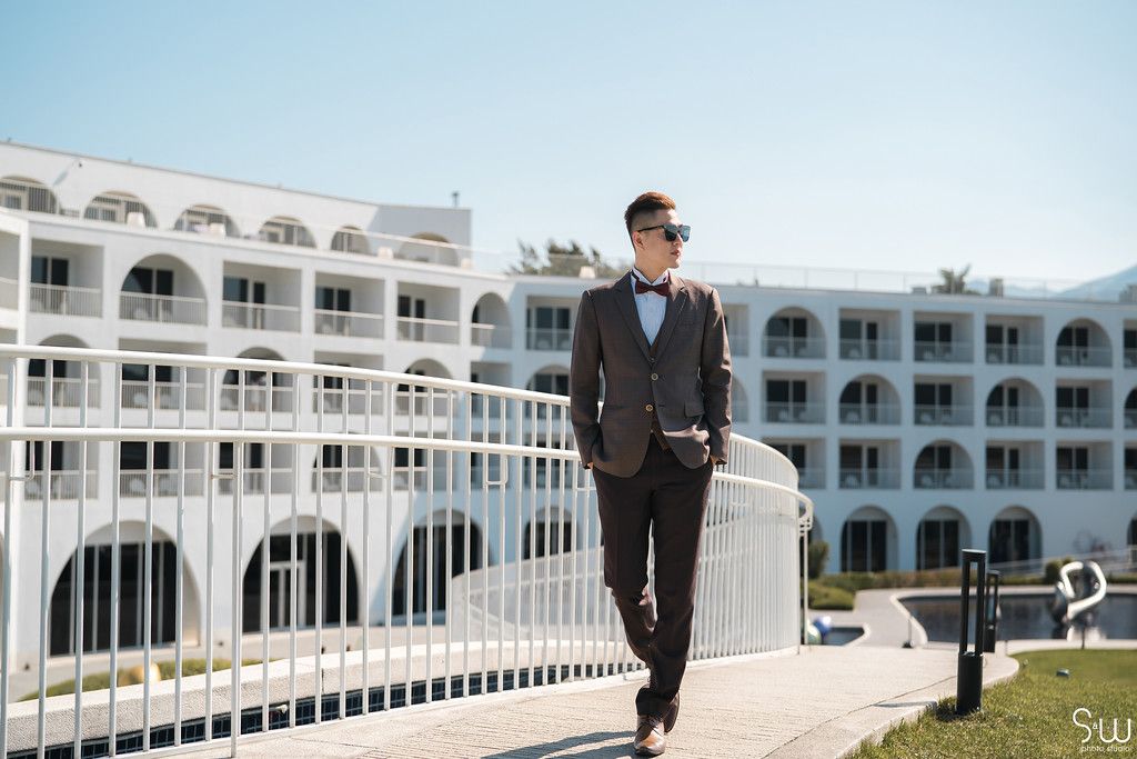 Groom in dark suit poses on white balcony with Mediterranean-style hotel in background.