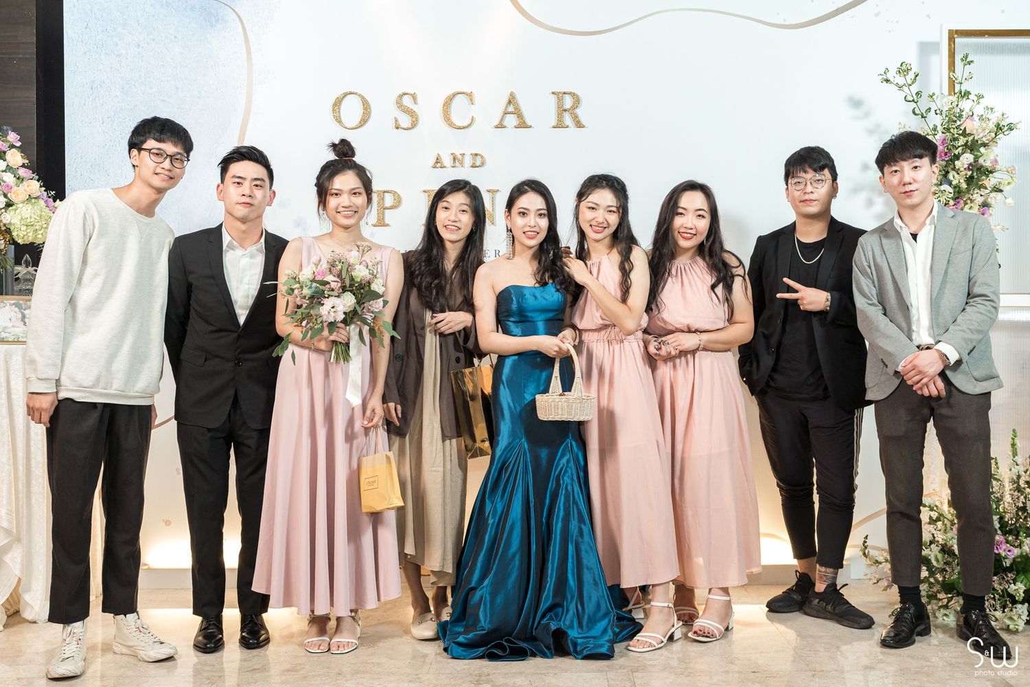 Group photo at a wedding with guests in formal attire standing in front of a floral backdrop with 'OSCAR' signage.