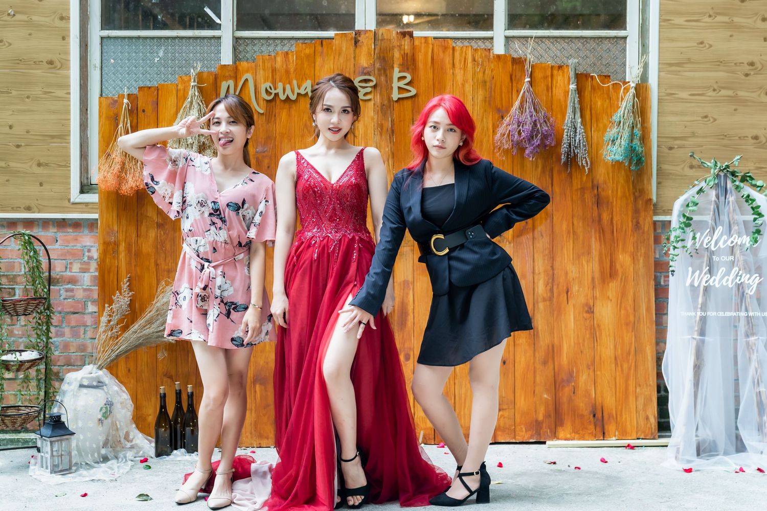 Three models in floral, red, and black dresses strike poses against a decorated wooden backdrop.