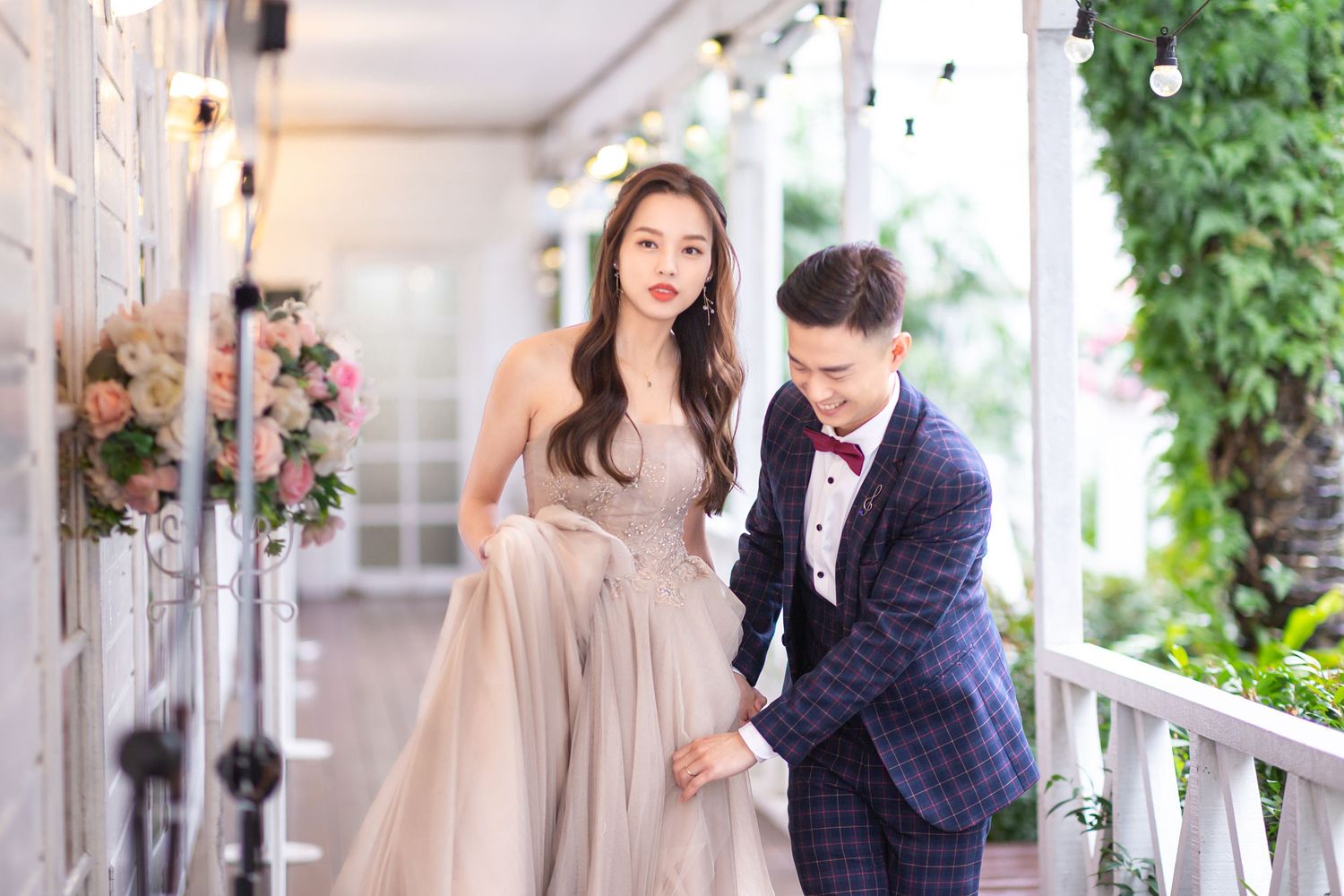A couple in formal wedding attire poses together on an elegant white porch decorated with floral arrangements.