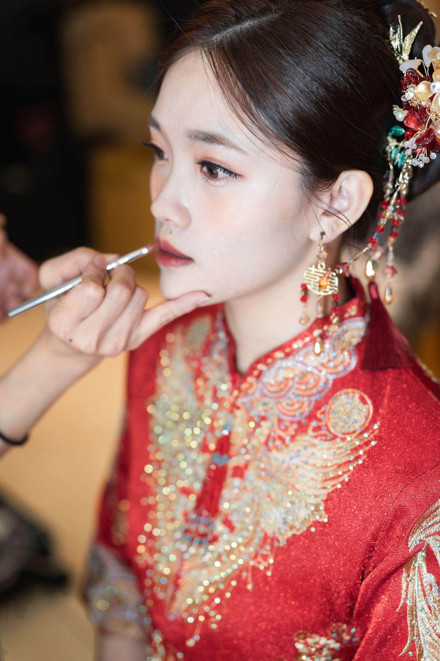 Makeup artist applies lipstick on bride wearing traditional red Chinese qipao dress with ornate gold embroidery and hair accessories.